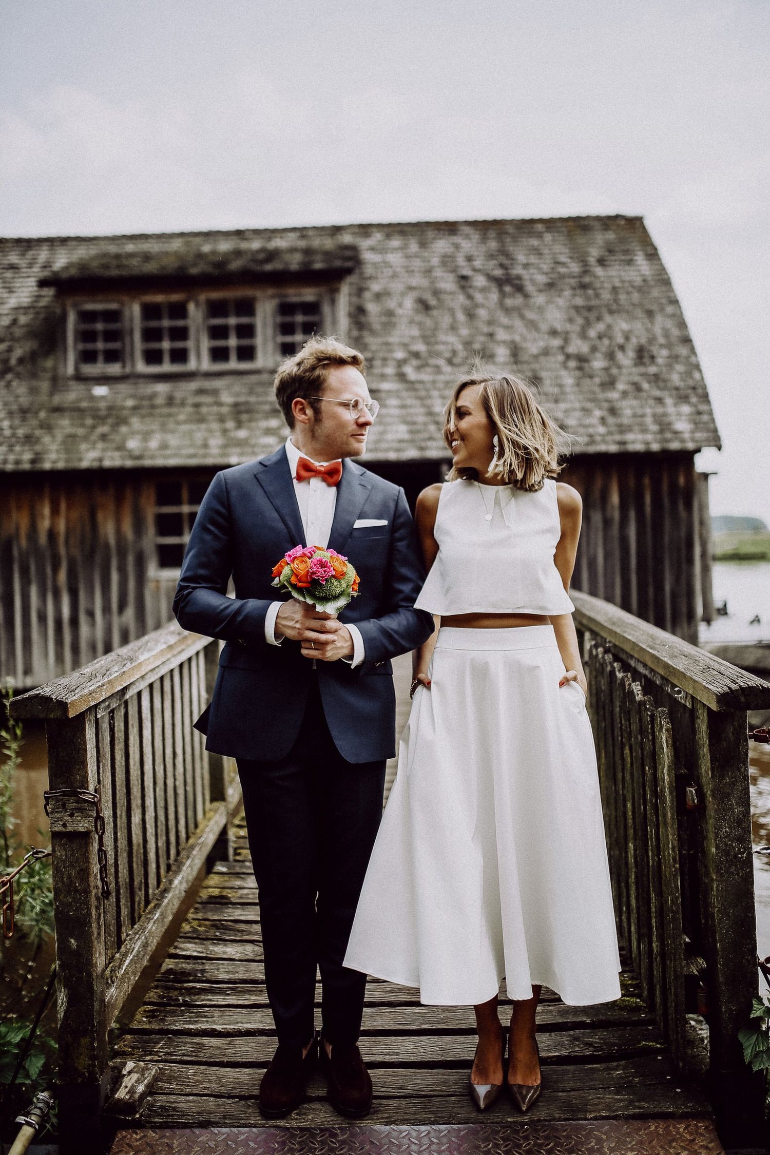 Couple in modern wedding attire standing on rustic wooden bridge with barn in background.