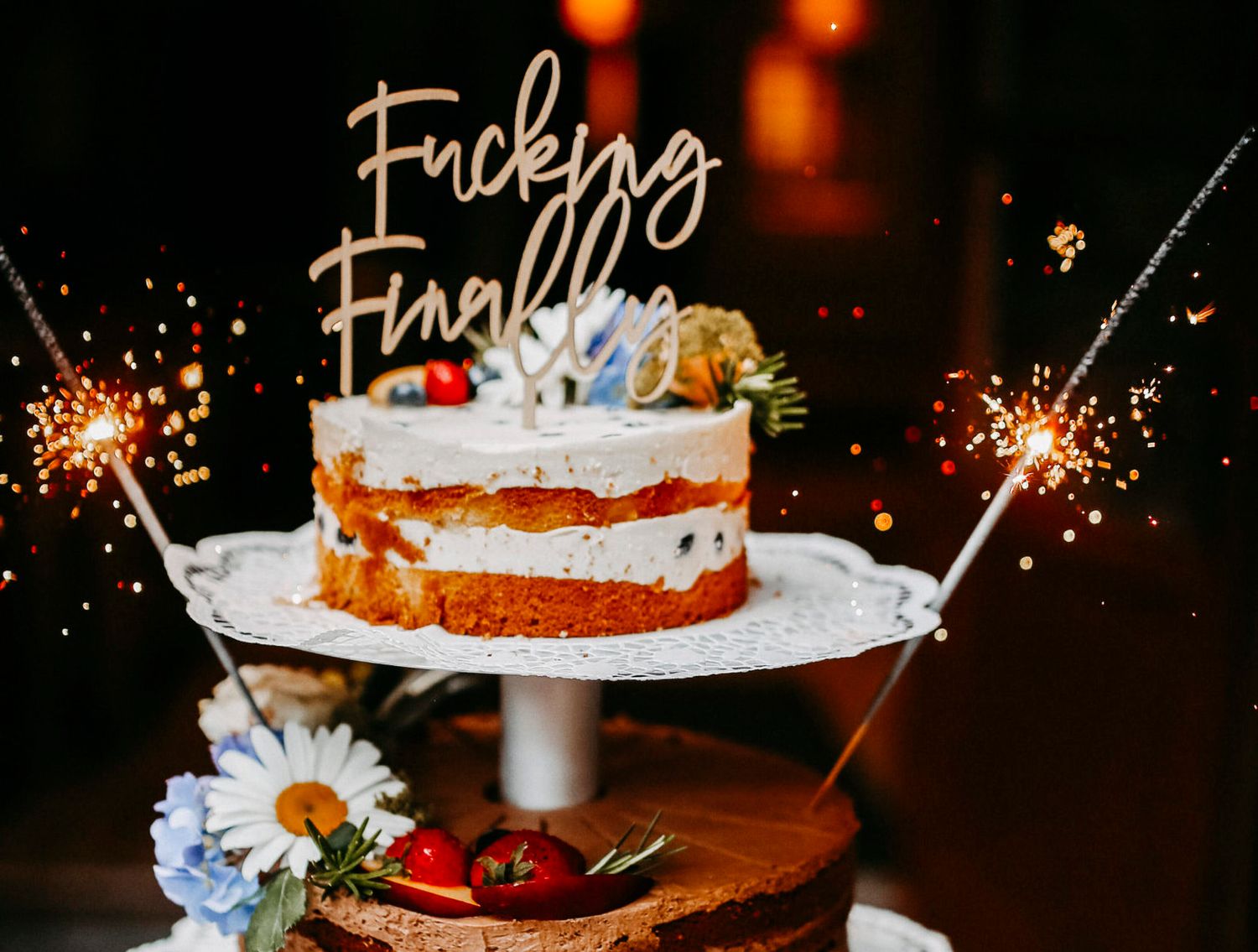 Festive wedding cake decorated with sparklers, daisies, and a cake topper on a stand with soft lighting.