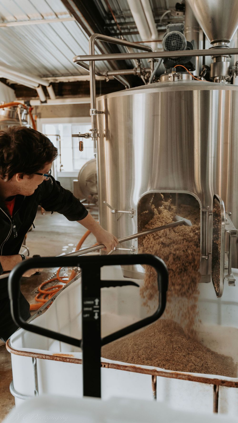 Worker operating large stainless steel brewery equipment with grains visible.