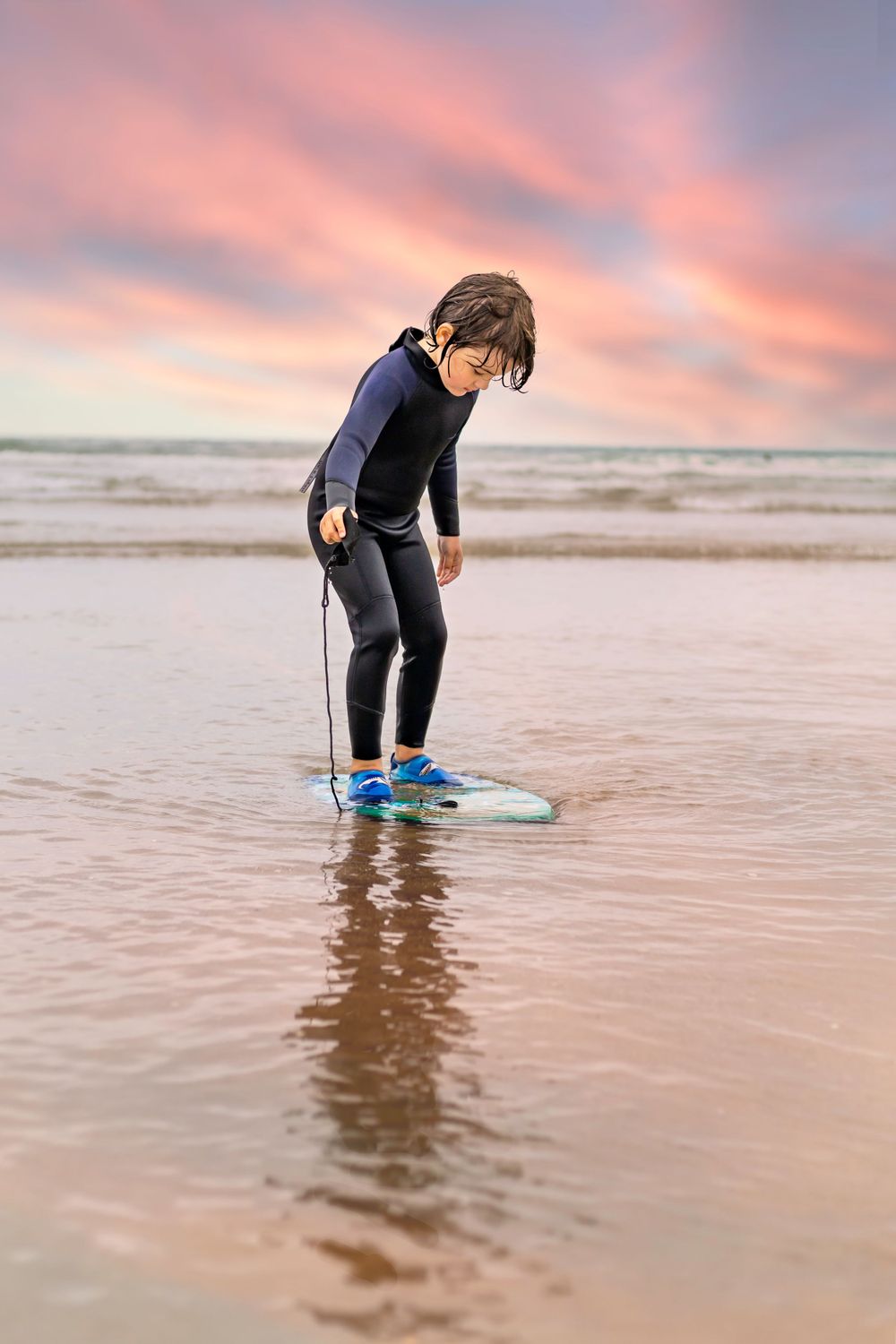 Young boy in a black wetsuit, stood on a small surf board looking down, shallow water ocean, pink sky behind