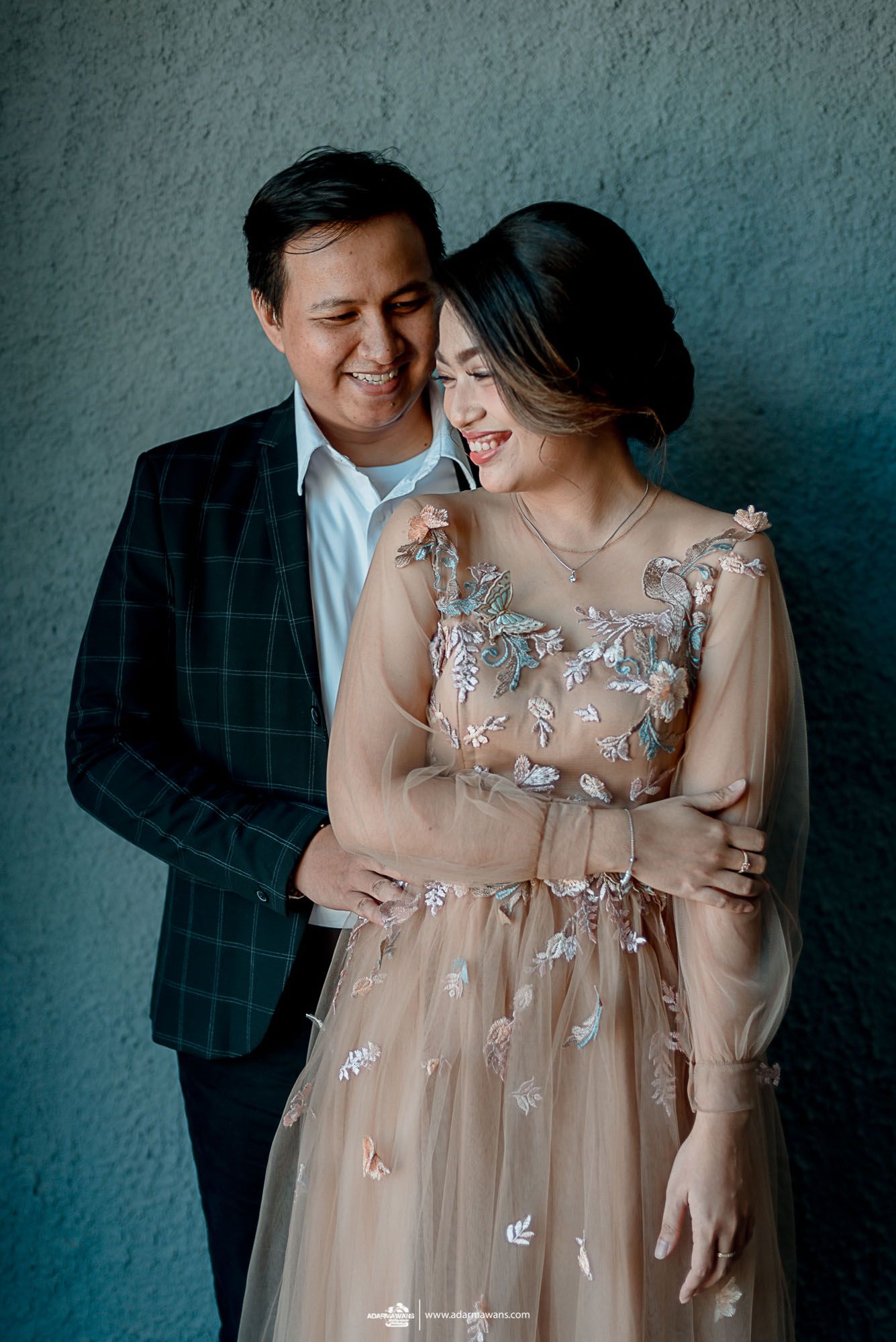 A romantic couple shares an intimate moment against a teal wall, with the woman wearing a delicate embellished tulle dress.