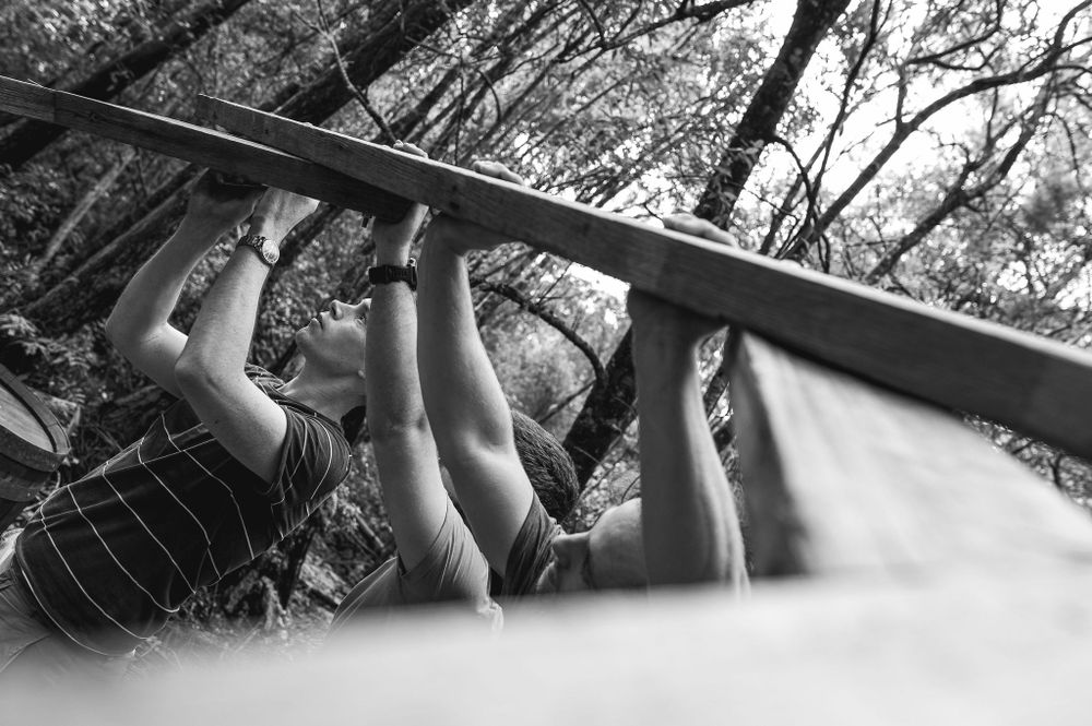 Black and white image of hands gripping and working on a wooden beam from below.