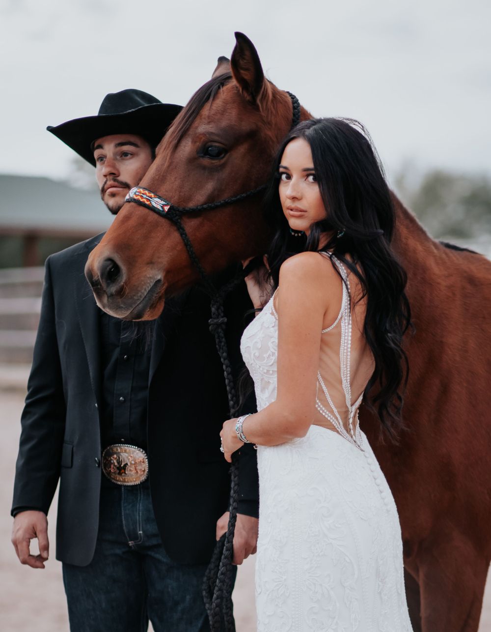 Romantic couple poses with a brown horse in outdoor wedding photos against a moody backdrop.