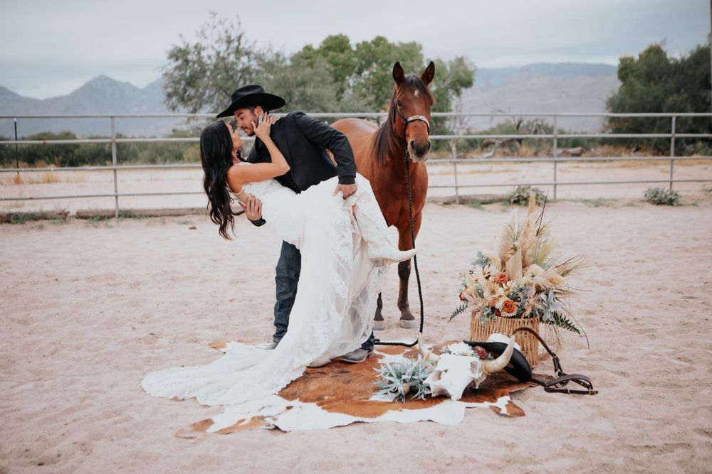 A bride in white dress poses with a brown horse in a desert ranch setting.