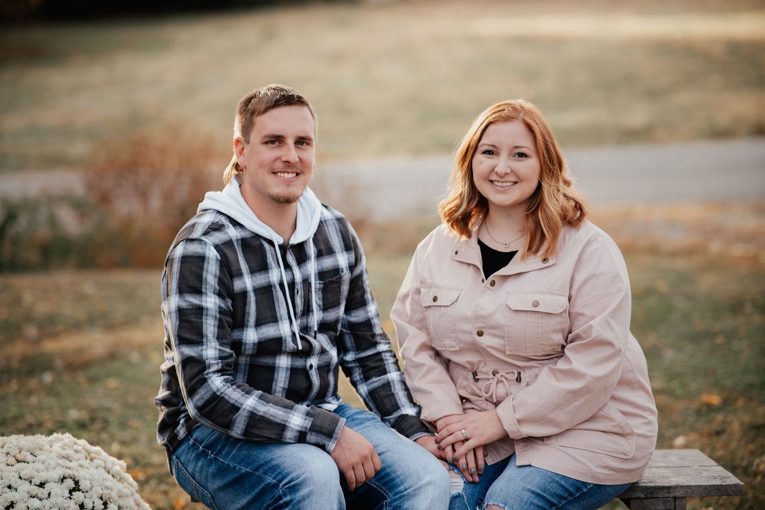 A couple sits together outdoors on a rock wearing casual sweaters in warm autumn lighting.