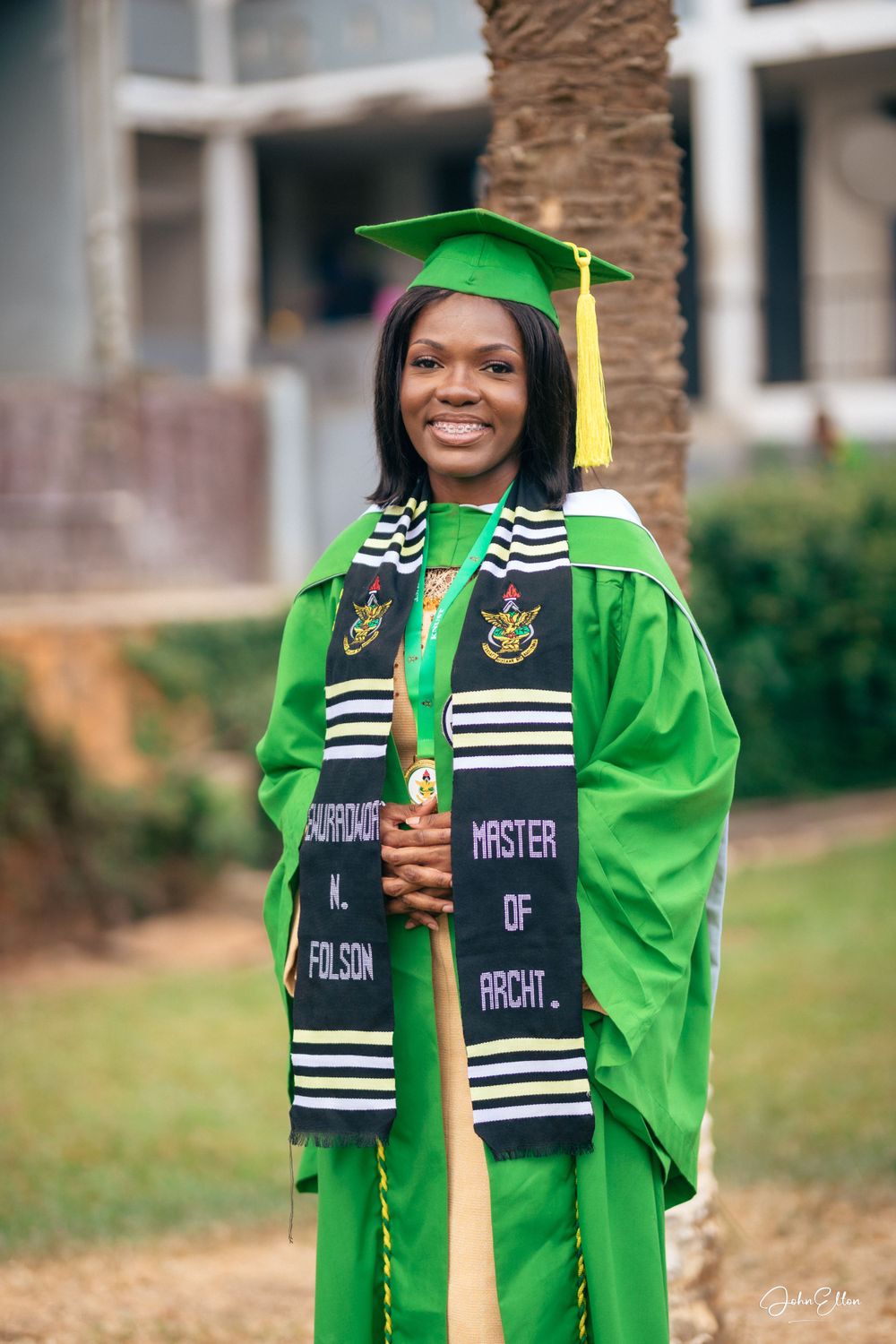 Graduate in bright green academic regalia and striped stole poses for celebratory photos outdoors.