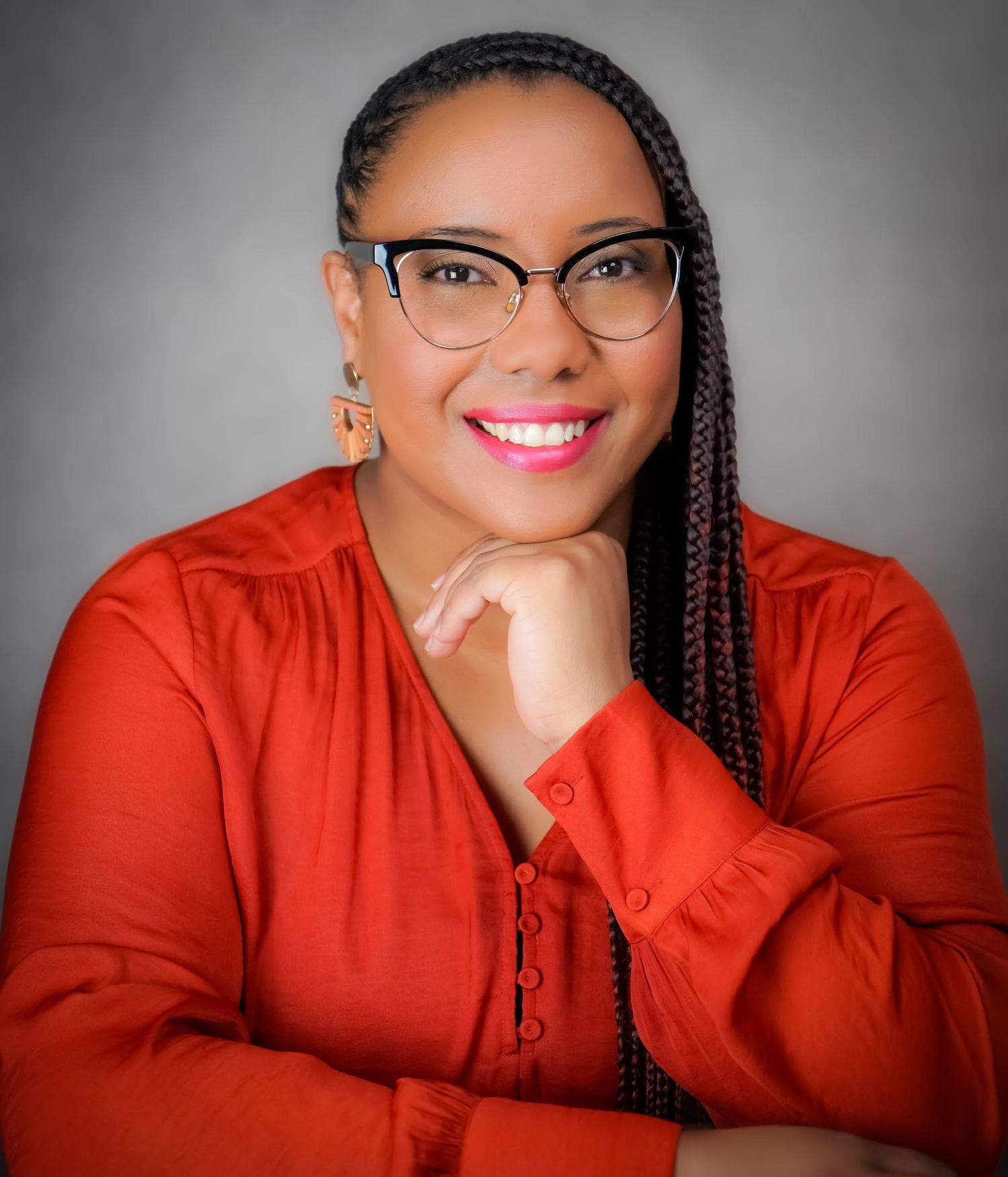 Professional headshot of a smiling woman in red blouse wearing glasses with braided hair.