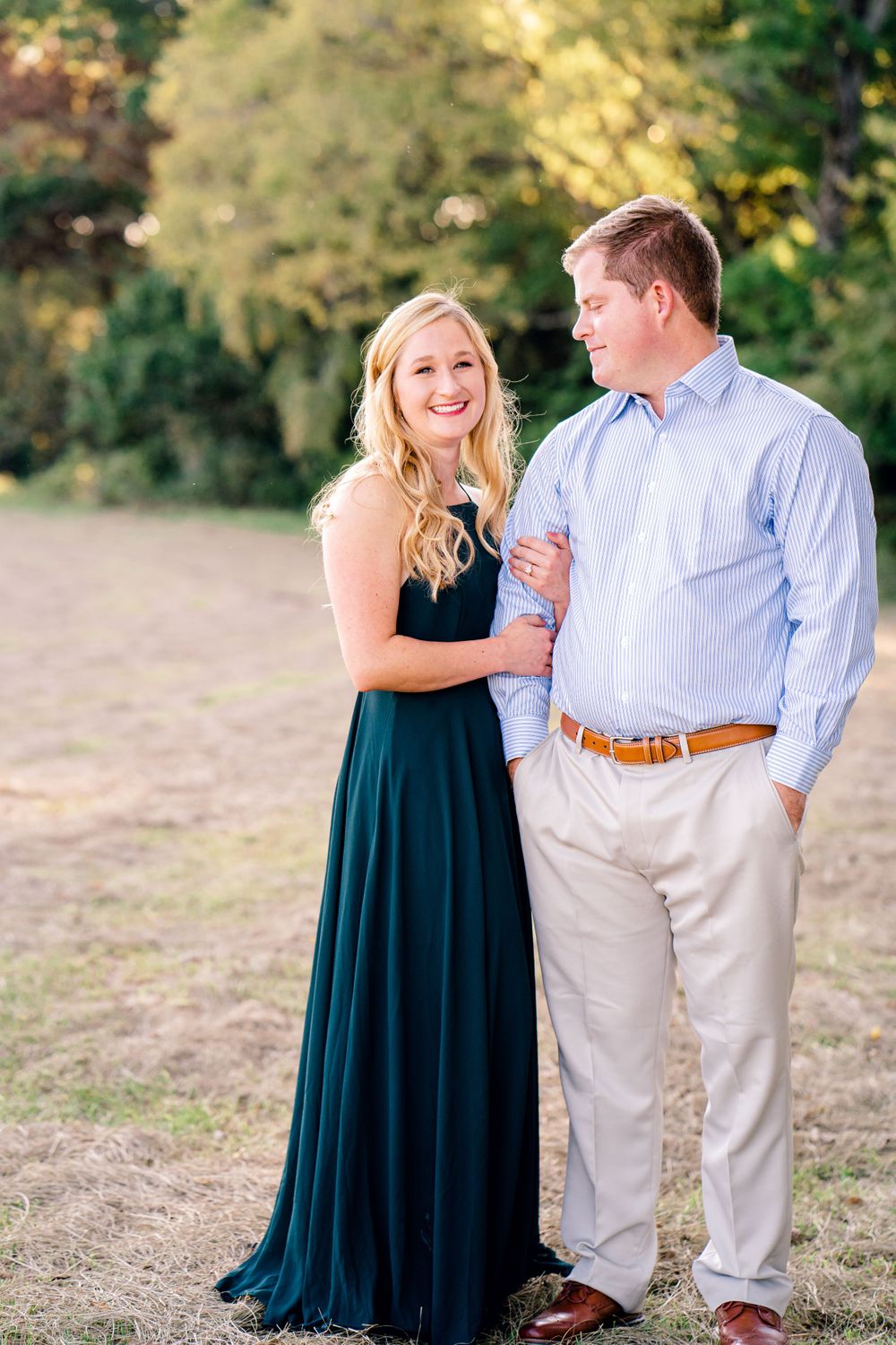 A couple stands together smiling in formal attire against a backdrop of autumn trees.