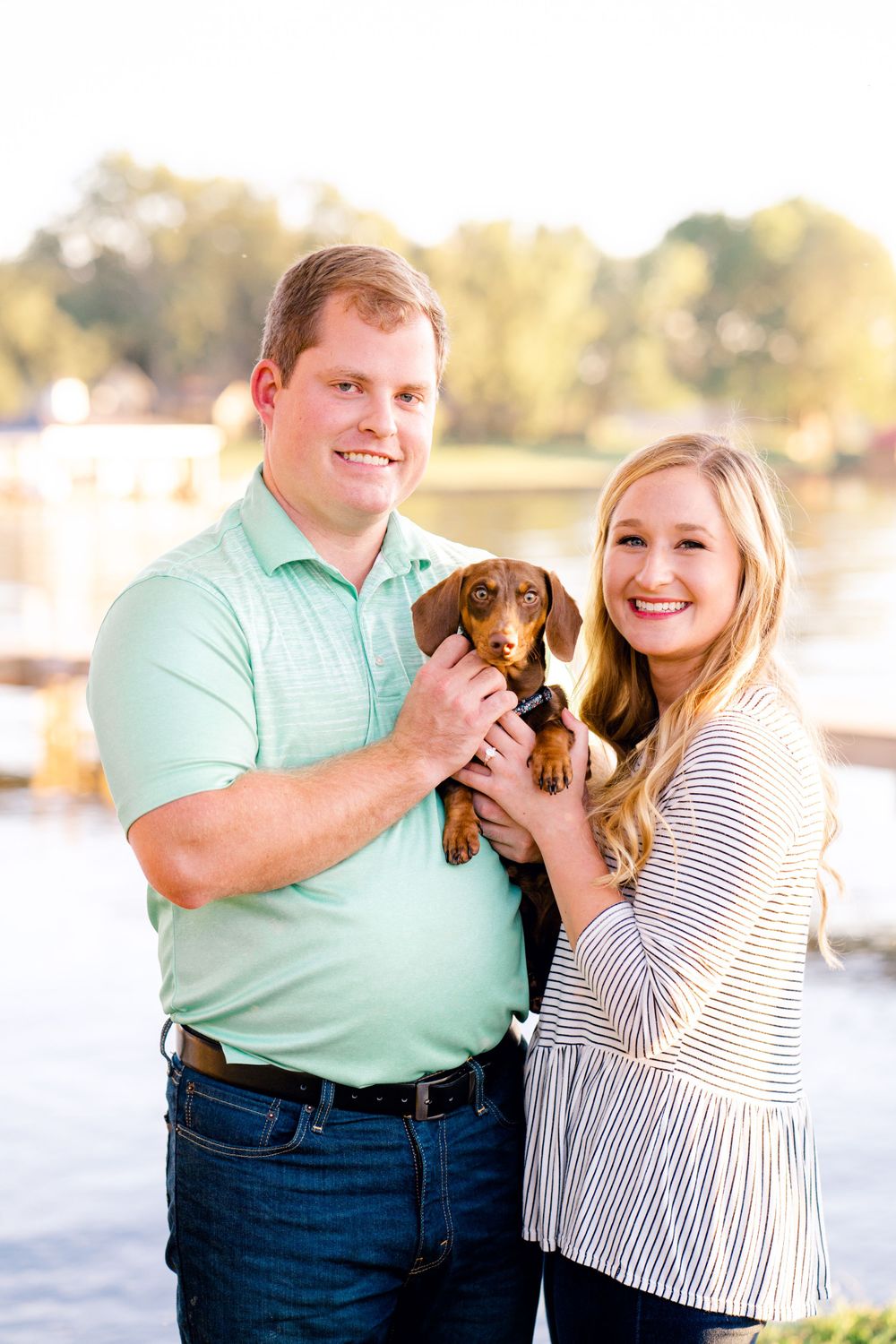 A couple poses outdoors holding their small dachshund puppy while wearing casual clothes near a waterfront setting.
