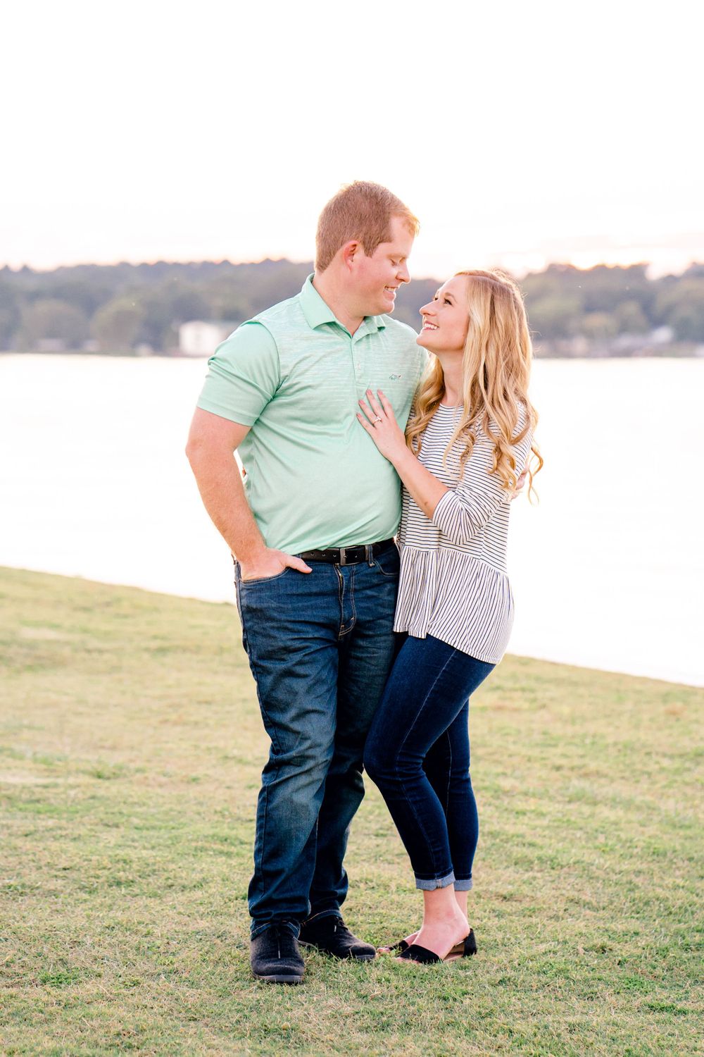 A romantic couple shares an intimate moment on grassy hillside by a lake during golden hour.