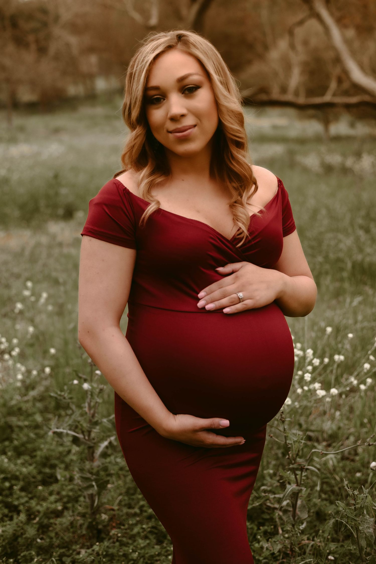 Elegant maternity photo of an expectant mother in a burgundy dress posing in a rustic outdoor setting.