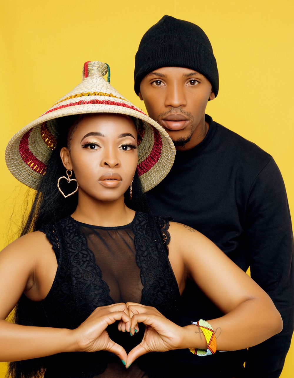 Stylish couple in black outfits posing against yellow background, making heart shape with hands.