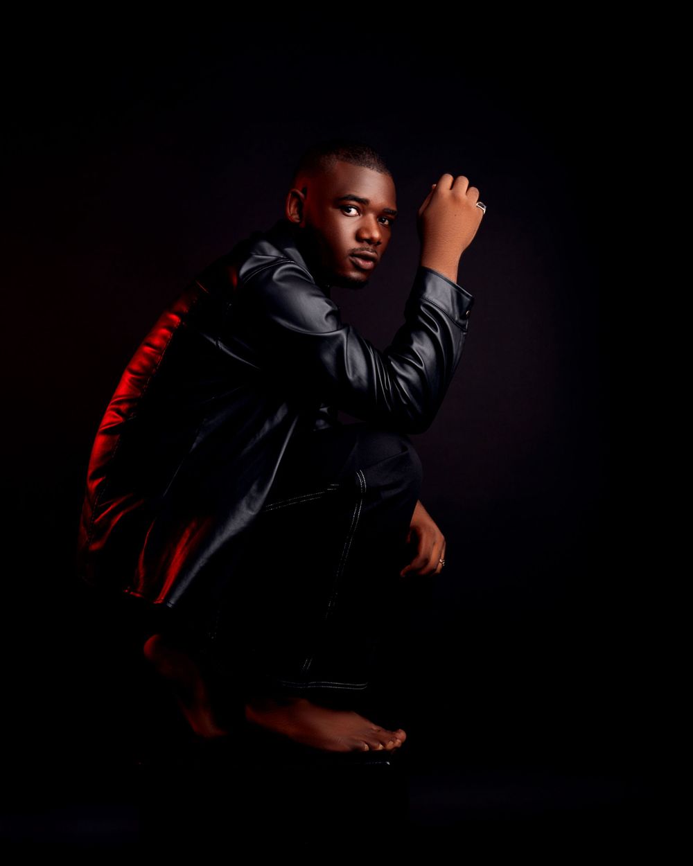 Dramatic portrait of person in black jacket crouching against dark background with red accent lighting.