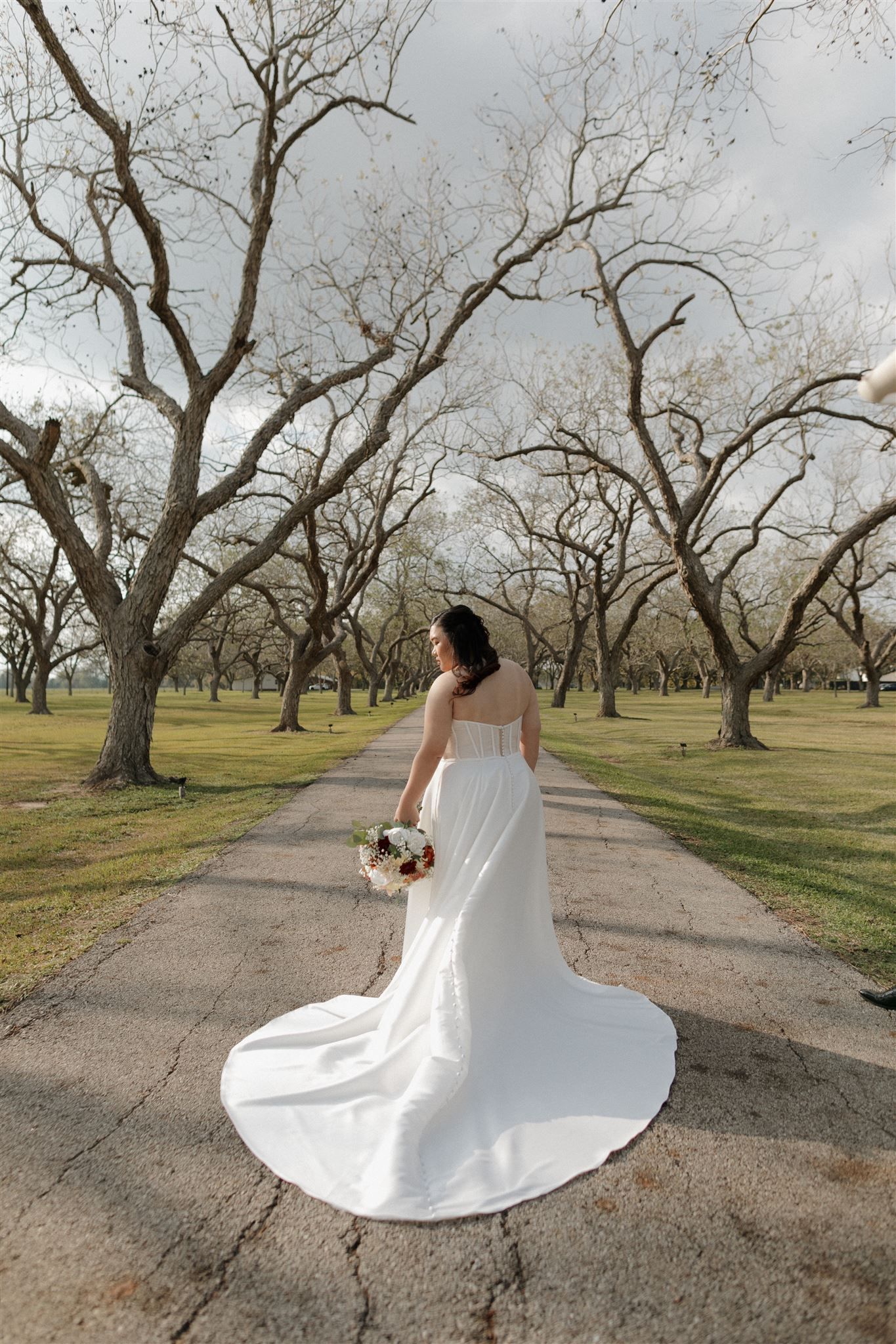 Bride in flowing white dress poses on pathway surrounded by bare trees.