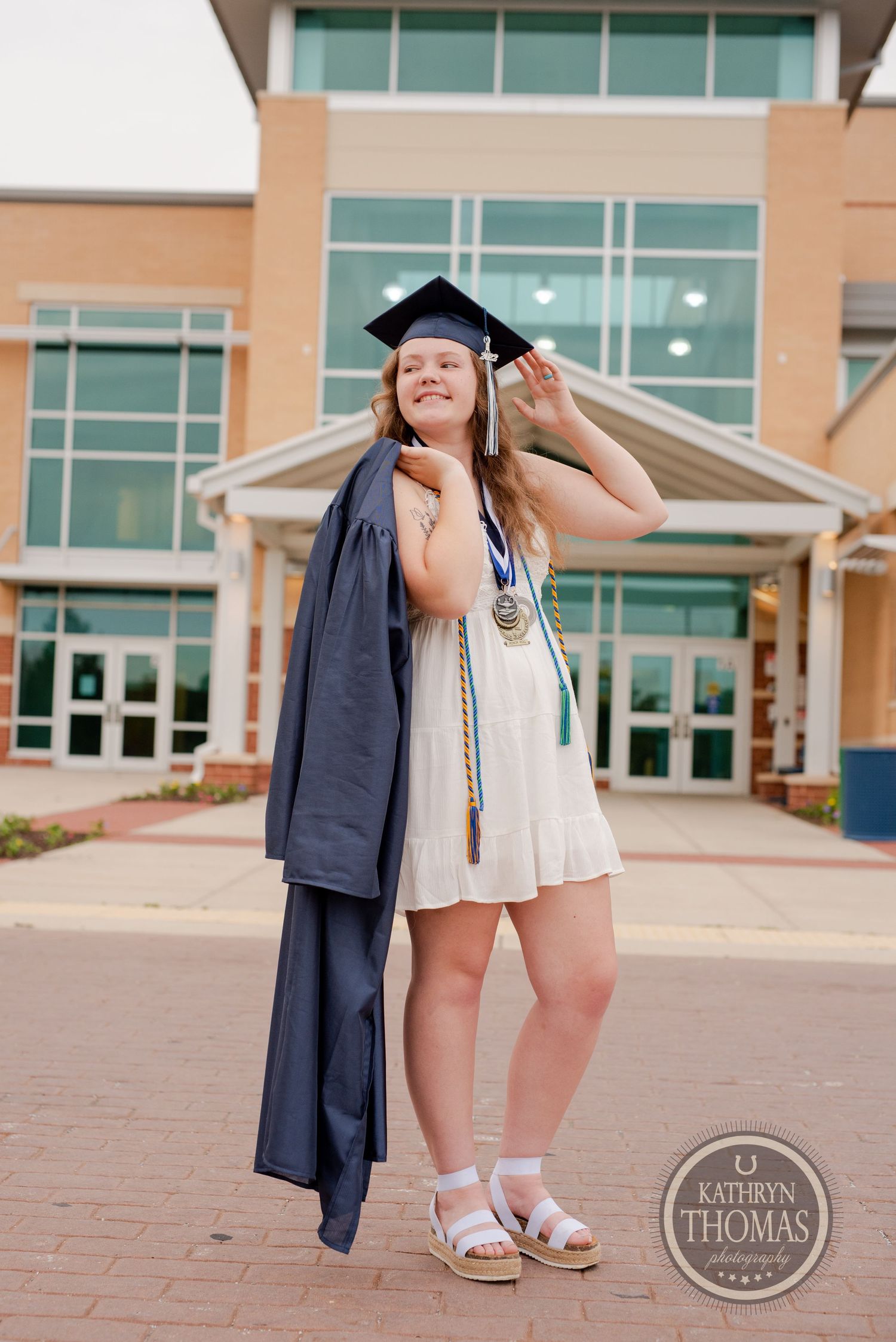 Graduate poses playfully in a white dress and navy cap and gown outside a modern school building entrance.