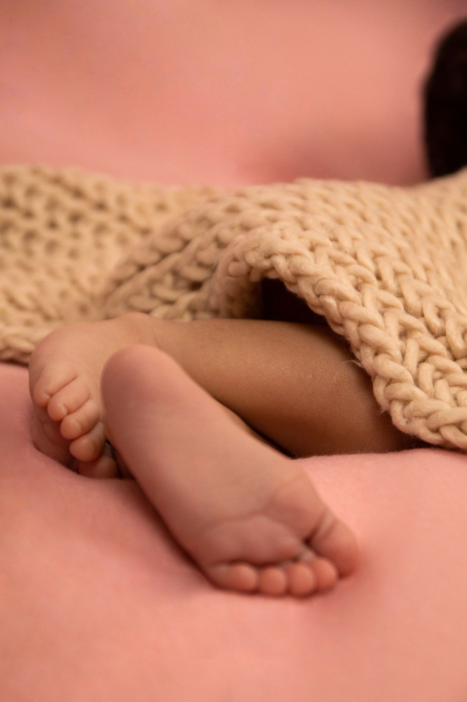 Tiny baby feet peeking out from under a chunky knit blanket on a pink background.