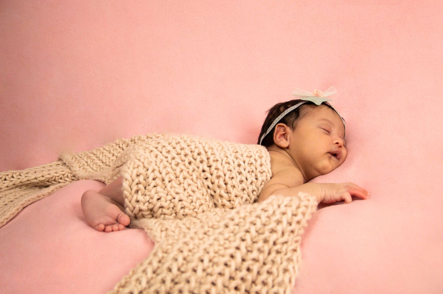 A sleeping newborn wrapped in cream knit fabric against a pink background.