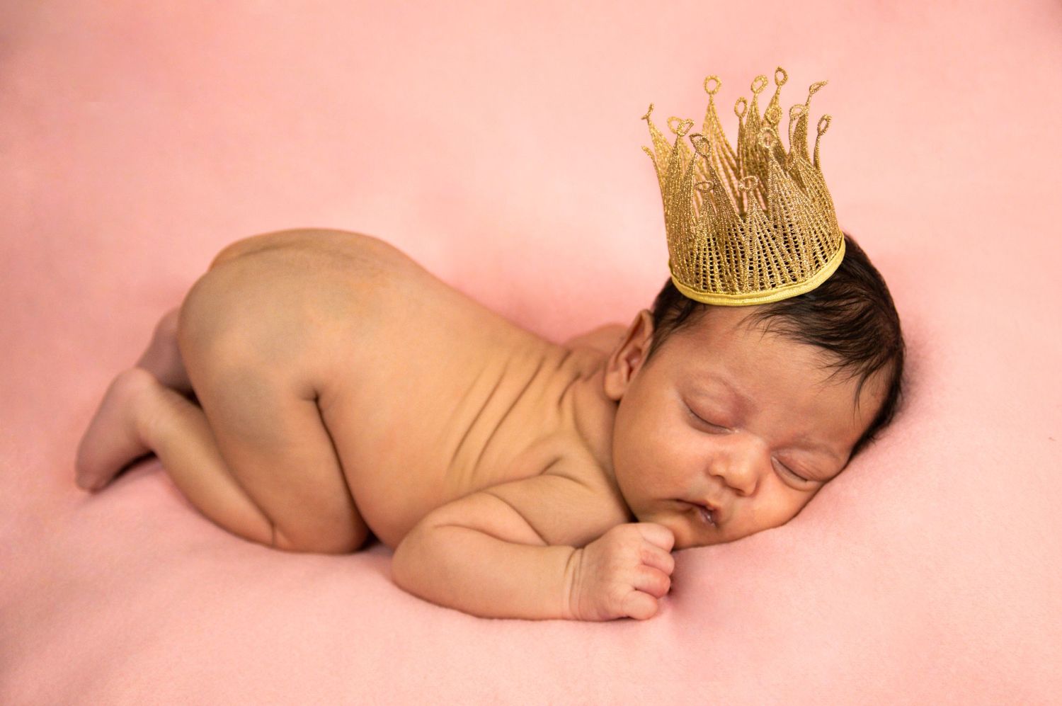 A sleeping newborn wearing a delicate gold crown on a pink background.