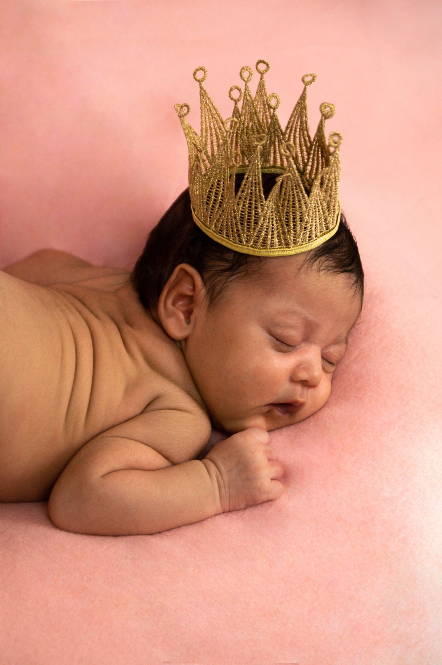 A newborn photography session featuring a sleeping baby wearing a golden crown against pink fabric.