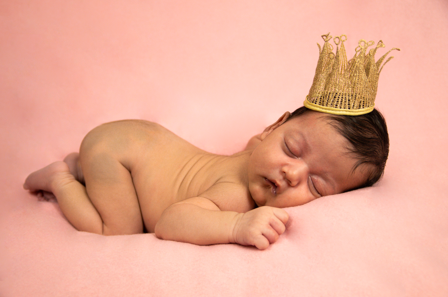A sleeping newborn wearing a gold crown during a professional photo session with pink backdrop.