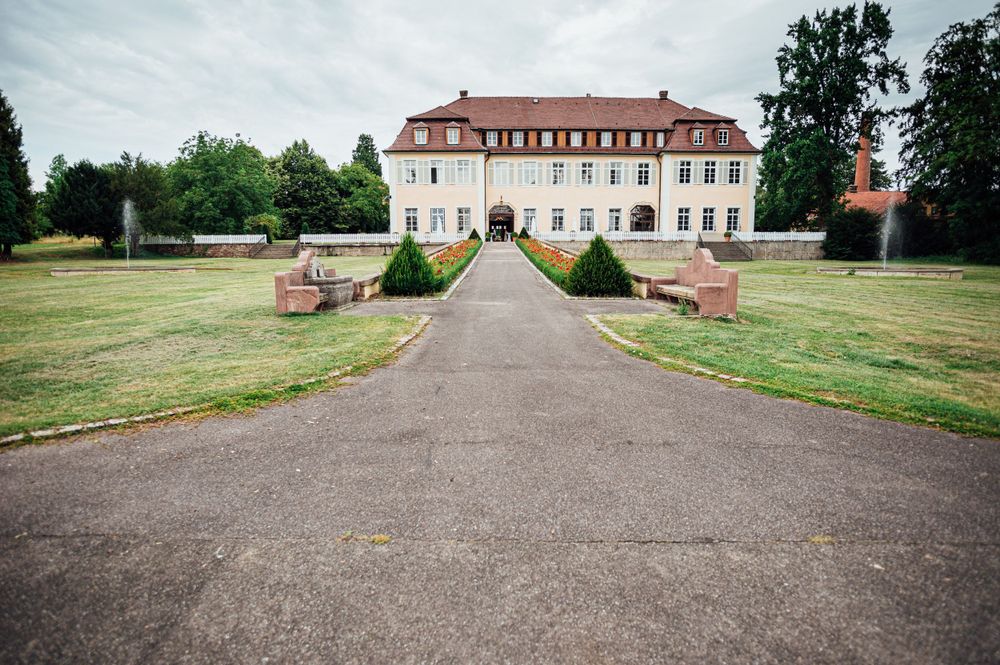 A grand historic mansion with red roof at the end of a long gravel driveway surrounded by manicured gardens.
