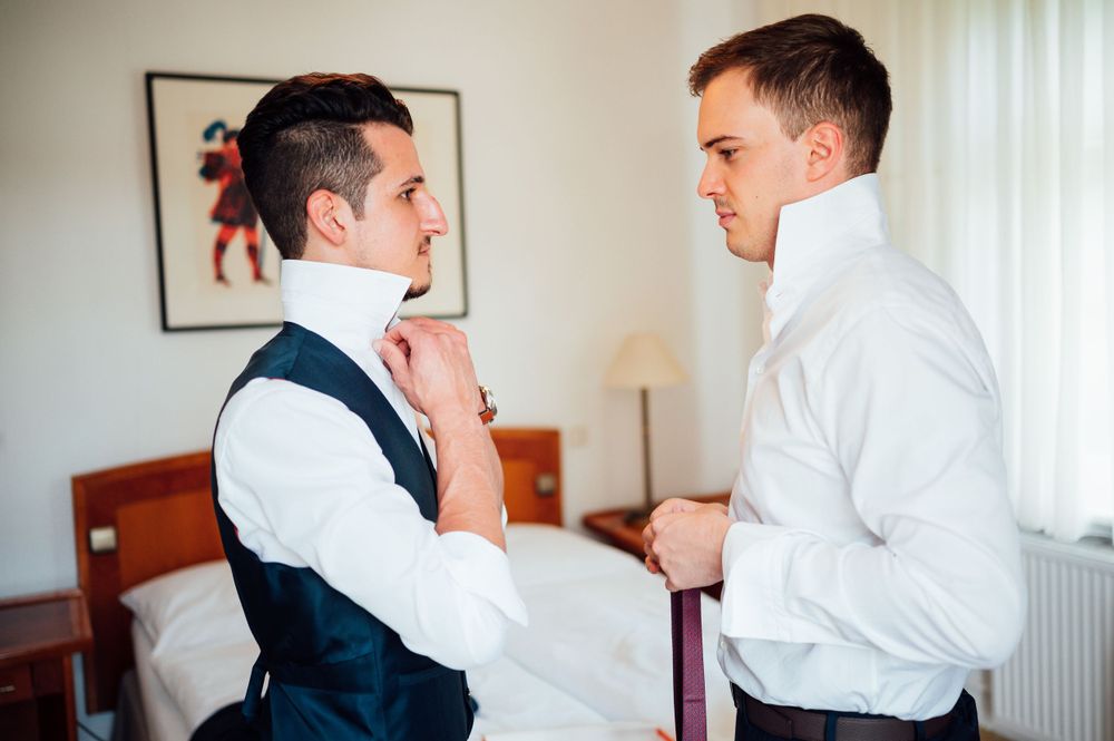 Two people preparing formal attire while discussing in hotel room with artwork on wall.