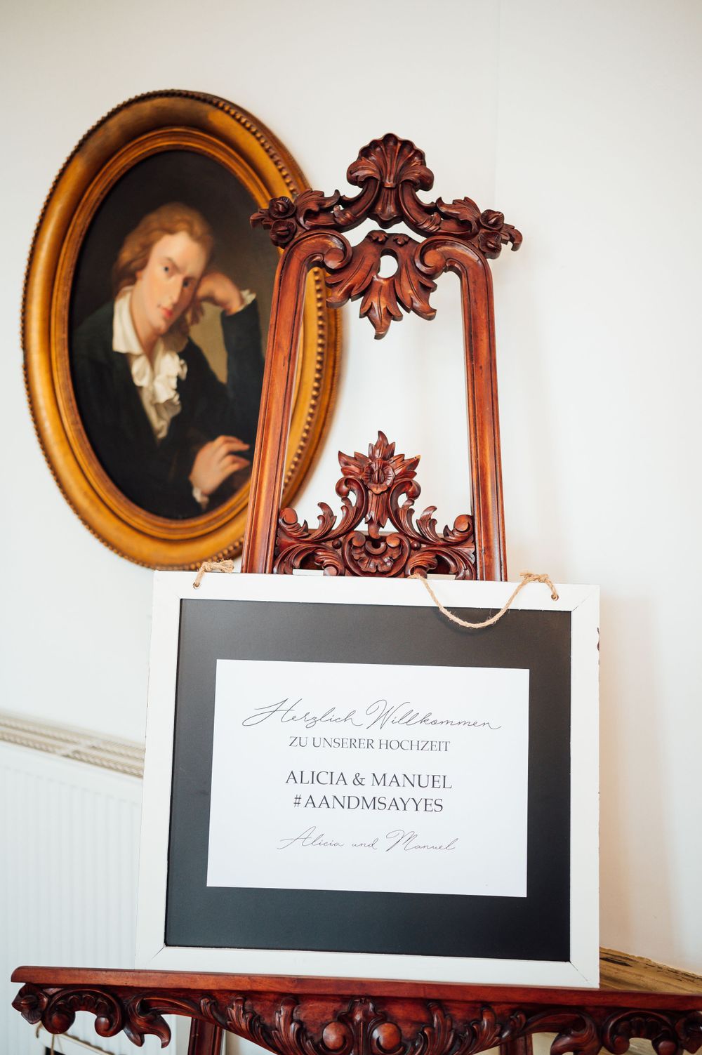 A vintage portrait painting in an ornate gold frame next to a black and white wedding invitation on an antique table.