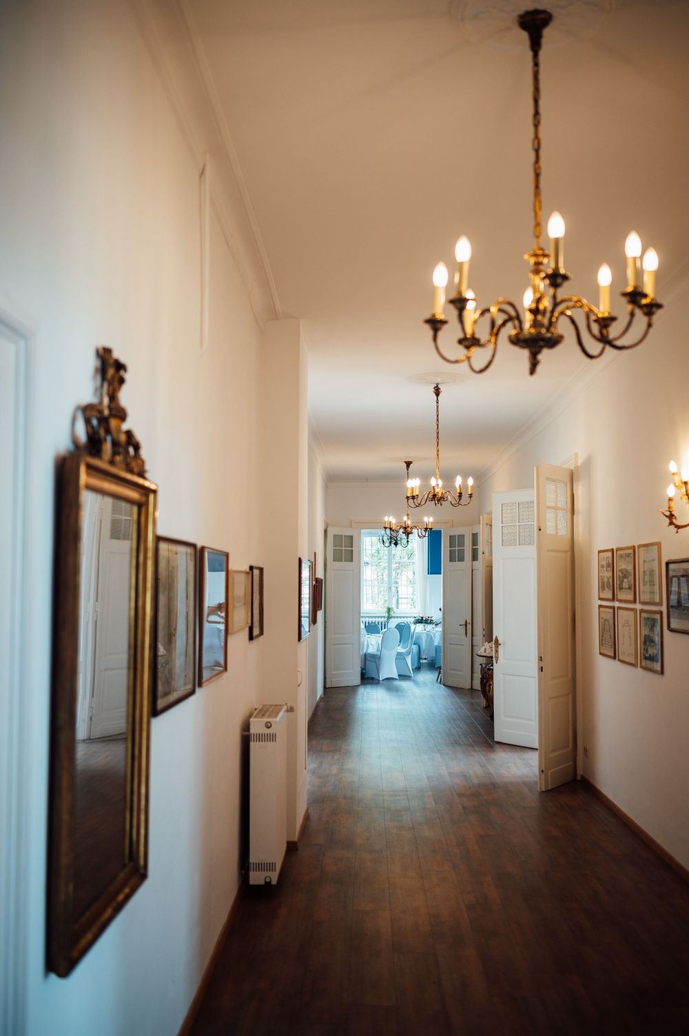 An elegant hallway with hardwood floors, white walls, and antique brass chandeliers leading to a bright room.