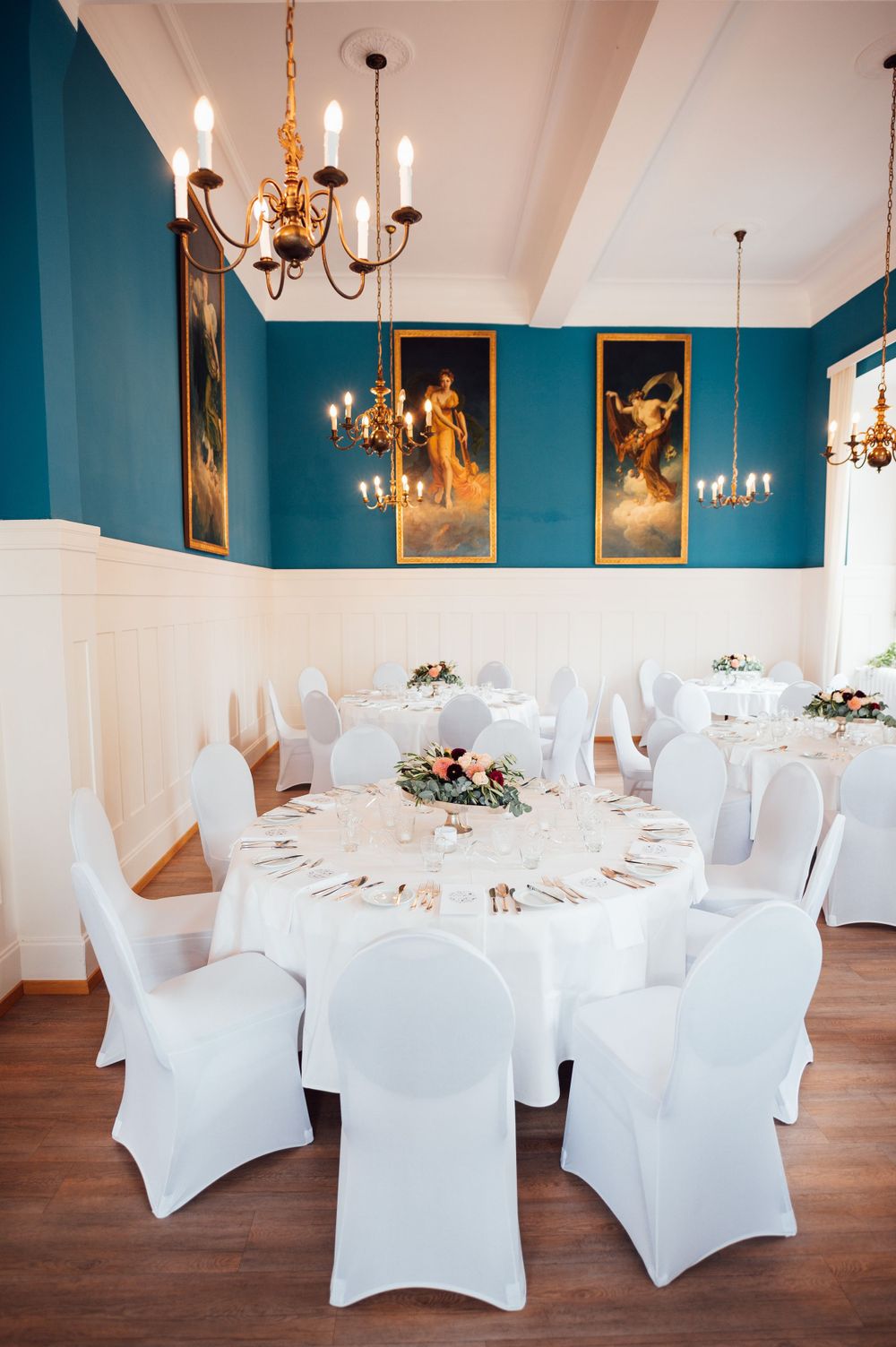 An elegant dining room with teal walls, crystal chandeliers and white-covered chairs around circular tables.