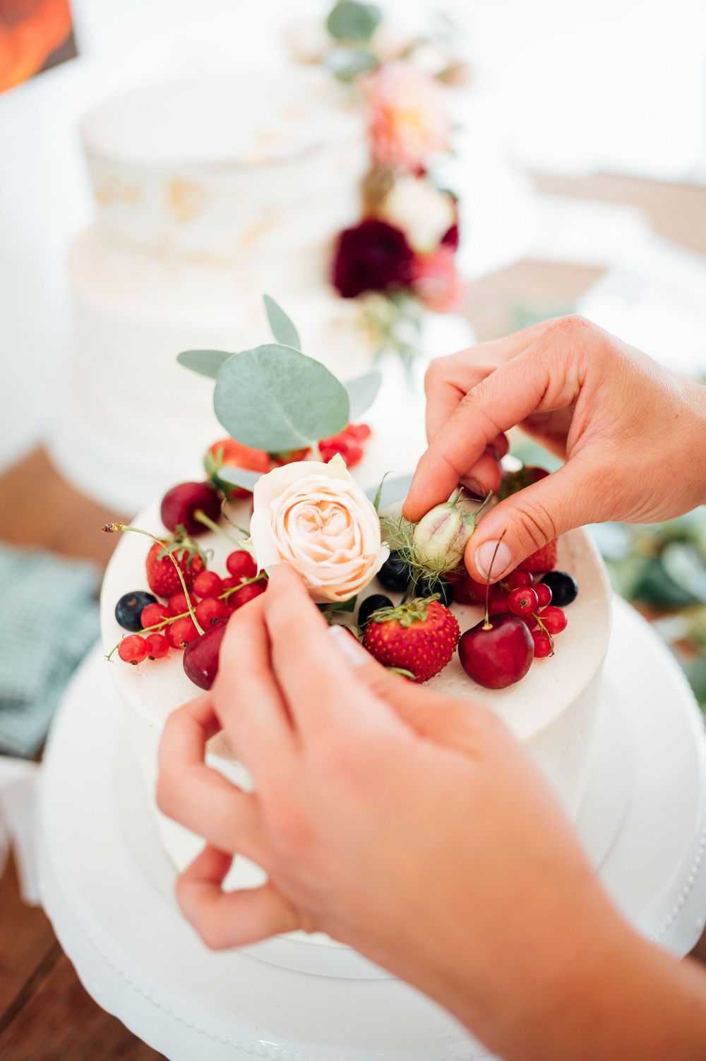 Hands arranging fresh berries, roses and strawberries on a white plate for an elegant dessert presentation.
