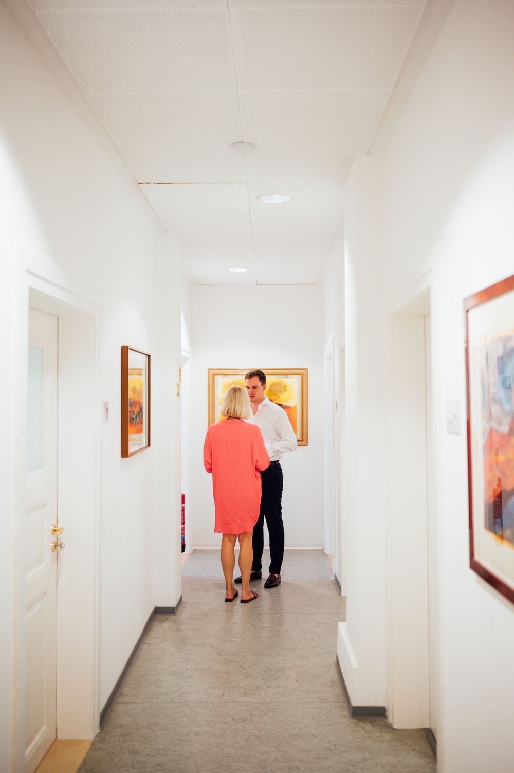 A bright white hallway with artwork displayed on the walls and a couple walking in the distance.