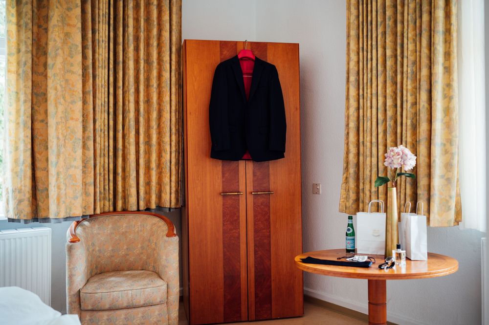 A navy suit jacket hanging in a wooden wardrobe beside a beige armchair and side table.