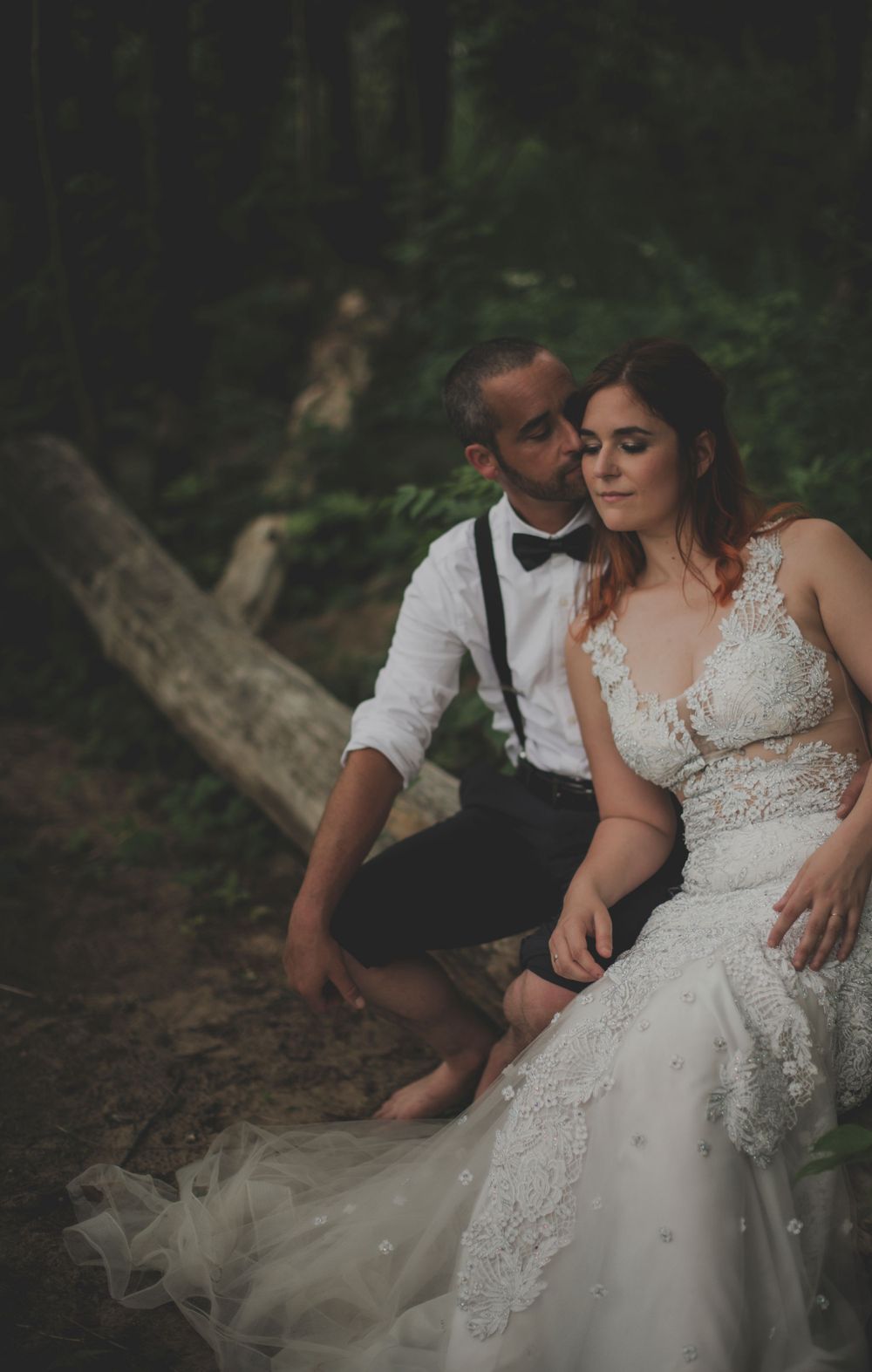 Newlywed couple in formal attire sitting intimately together in a woodsy outdoor setting.