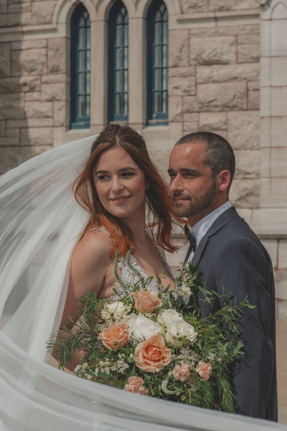 Bride and groom share a romantic moment outside a historic stone church with flowing veil.