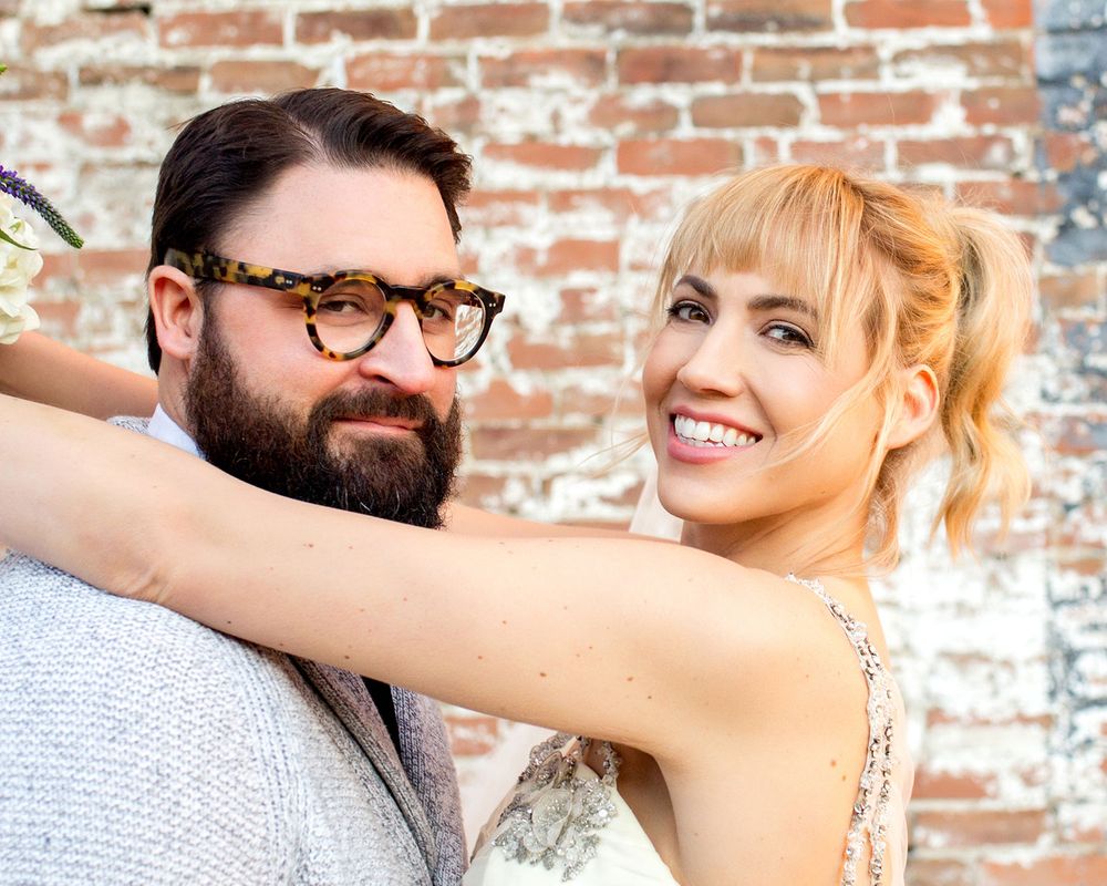 Romantic wedding couple shares intimate moment against weathered brick backdrop.