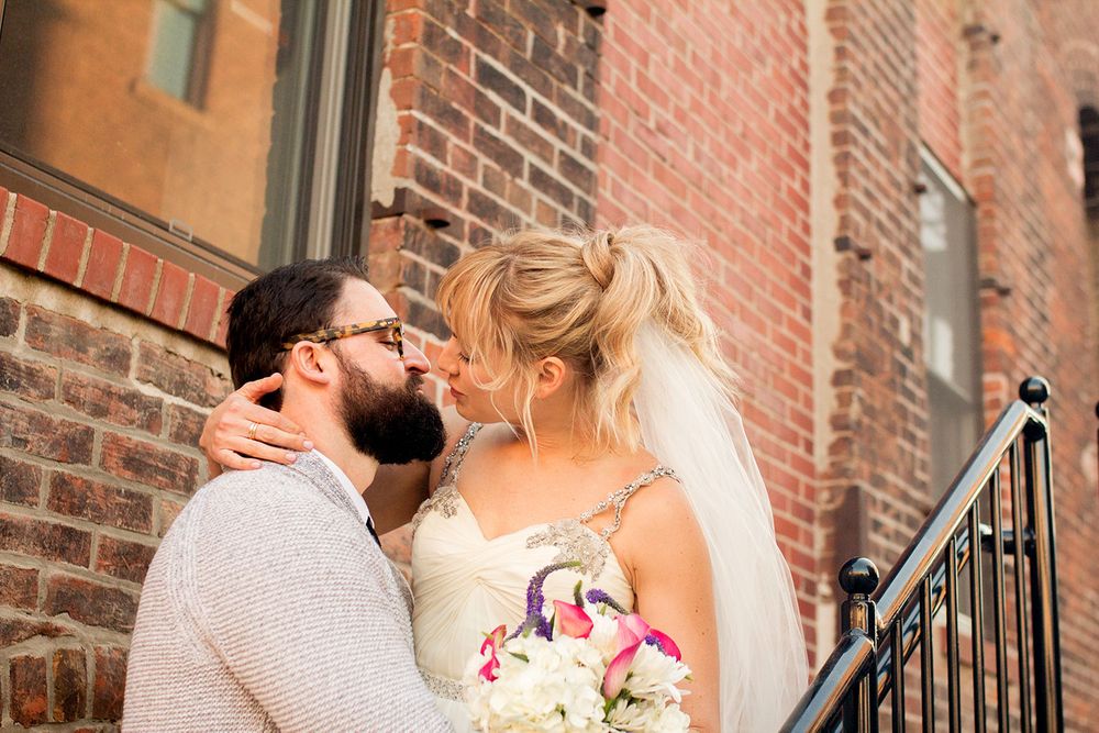 Romantic wedding couple sharing a kiss against a brick wall backdrop with flowing veil.