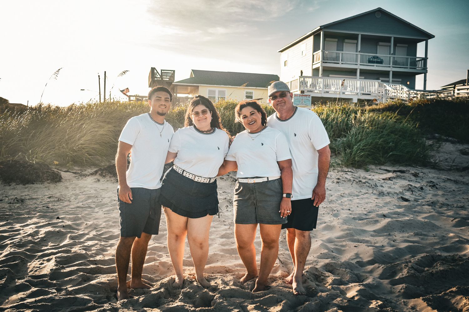 Friends in matching white shirts pose together at sunset near a beach house.