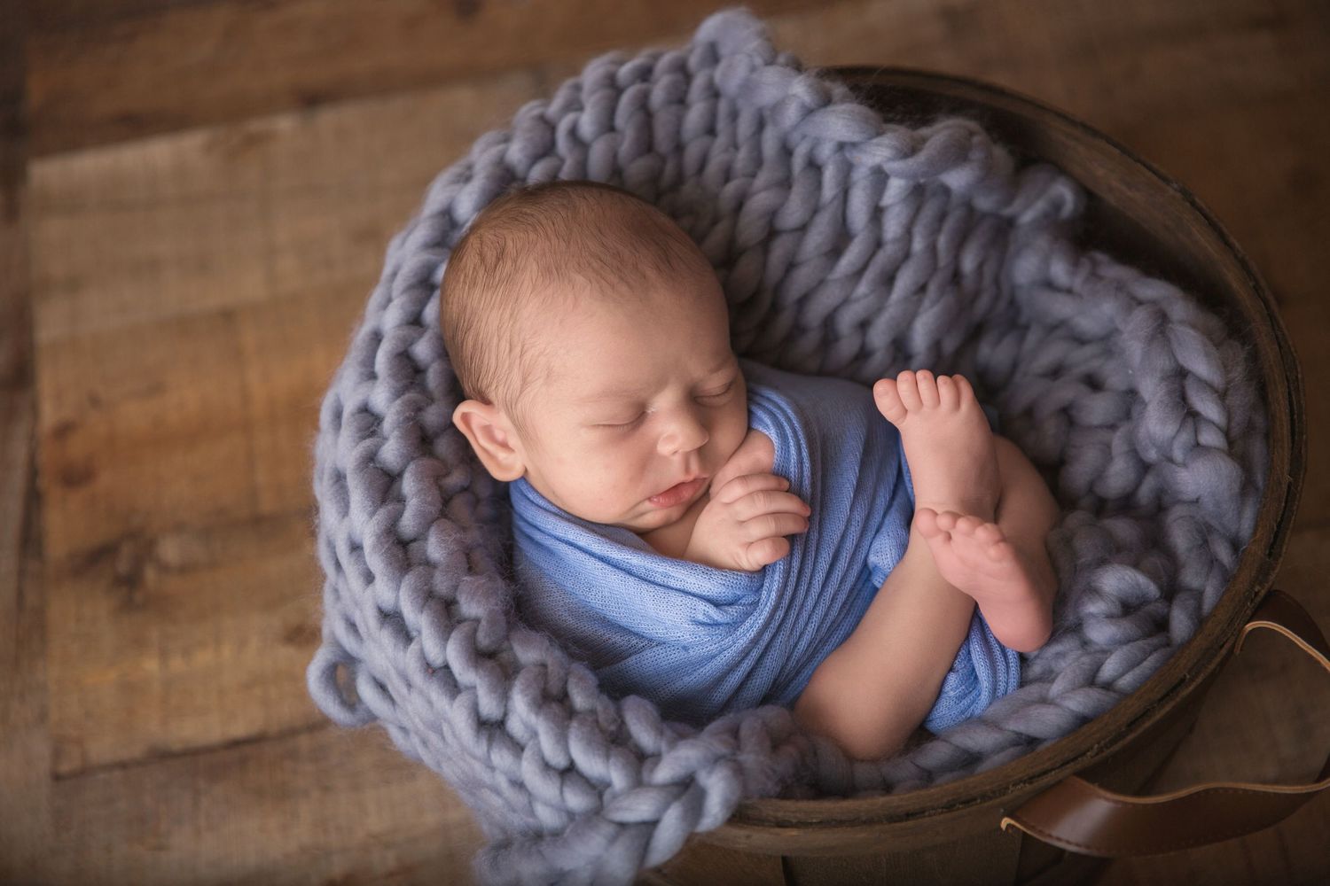 Sleeping newborn wrapped in chunky knit blue blanket on wooden backdrop.
