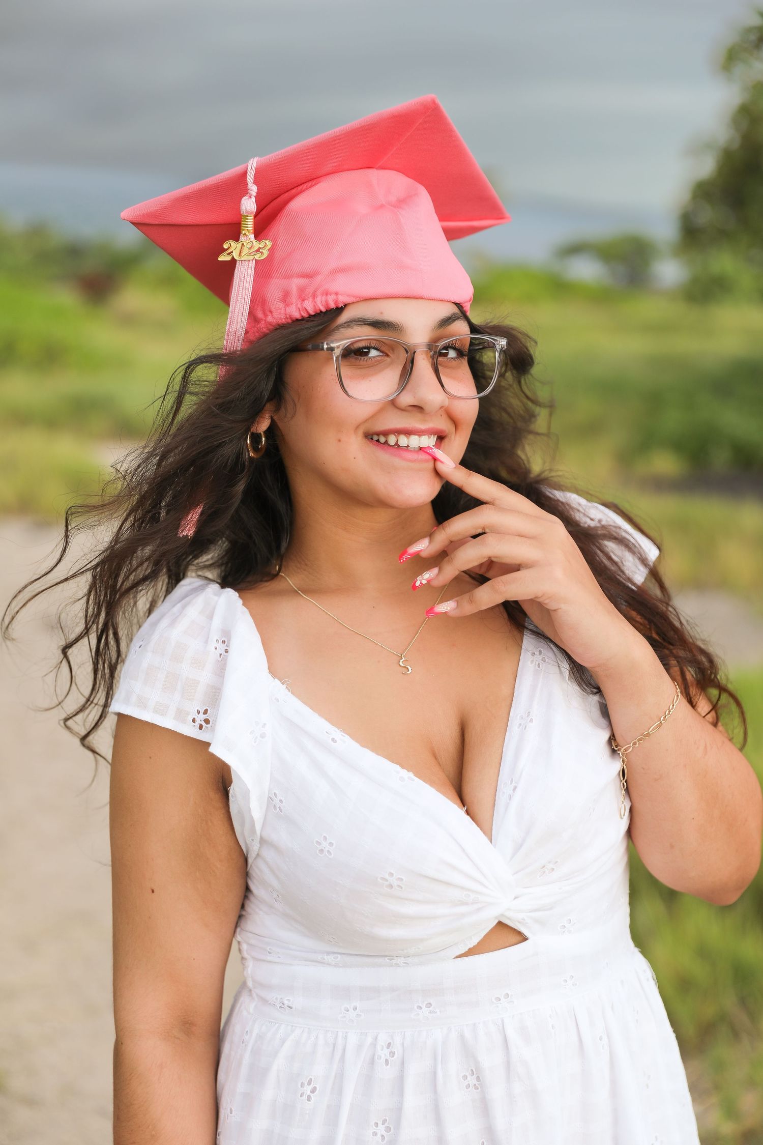 Graduate in white dress and pink cap smiles while wearing graduation attire outdoors.