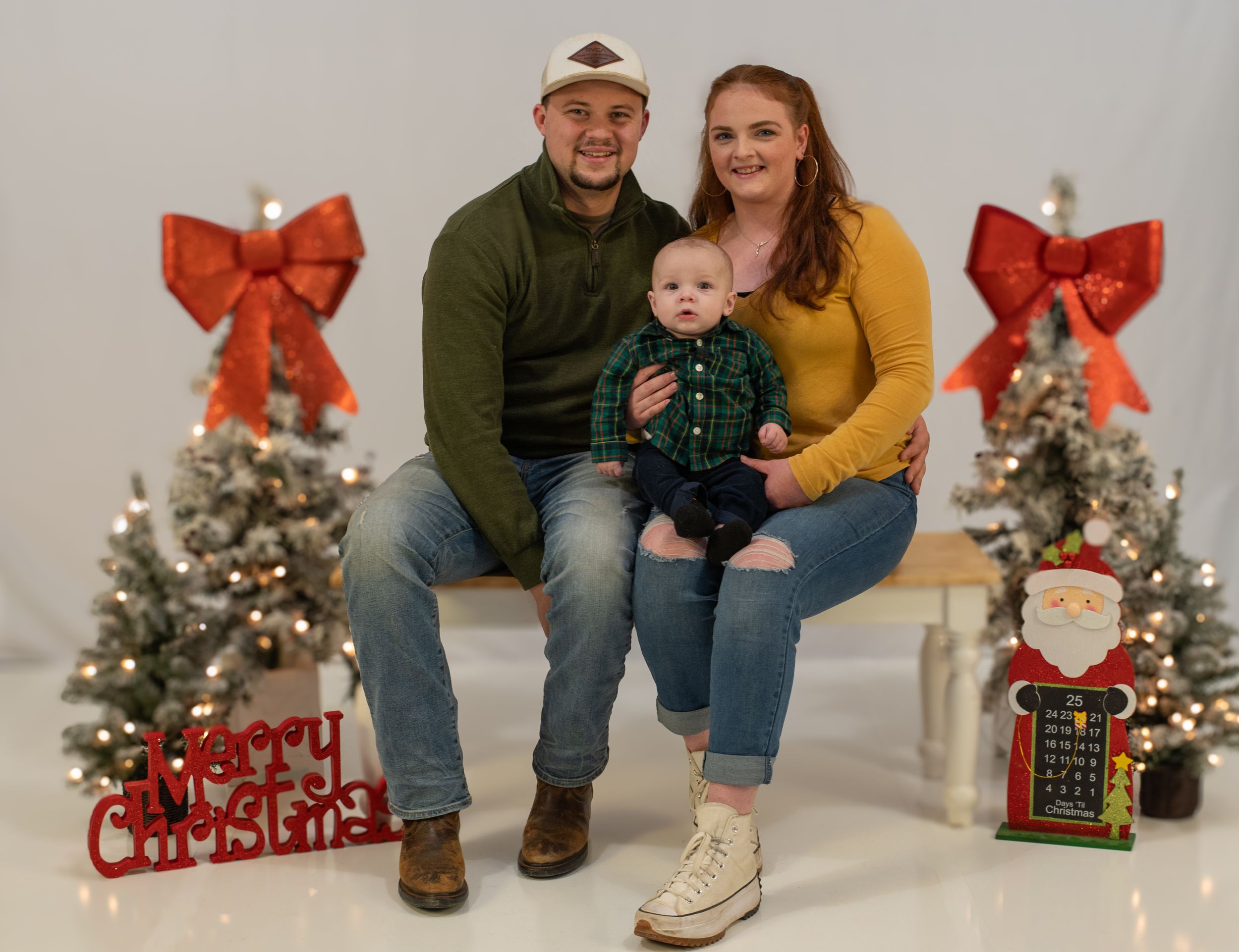 Family of three poses for Christmas photo with decorated trees and red bows in background.