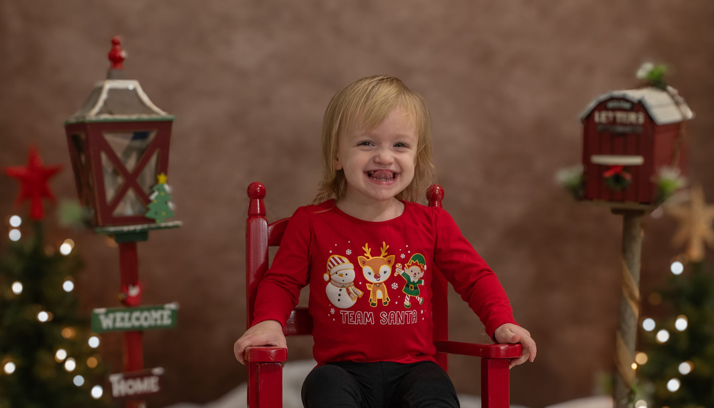 Smiling child in red holiday sweater sits in red chair with Christmas decorations in background.