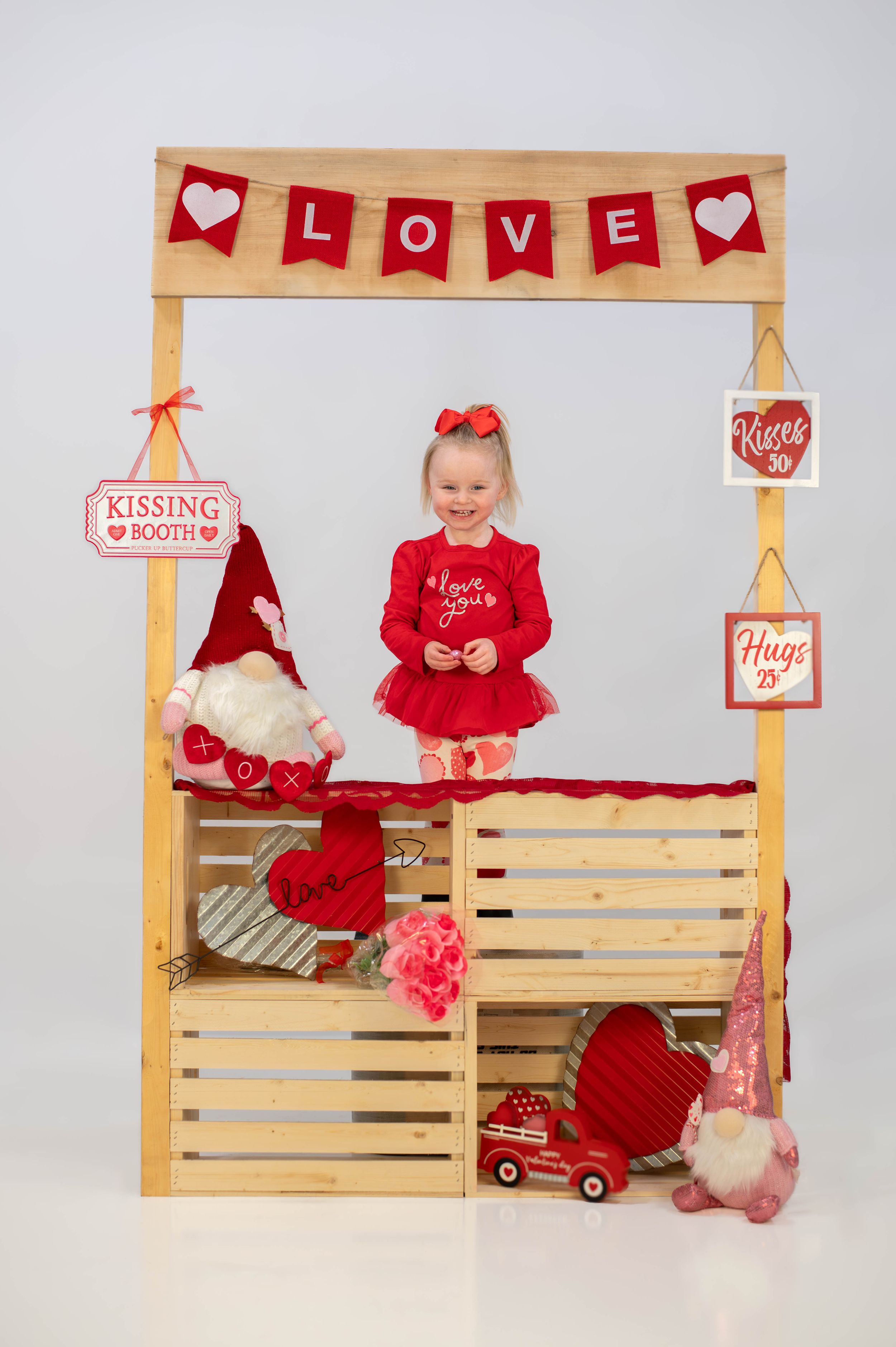 Child in red outfit poses at Valentine's themed photo booth with wooden crates and decorations.