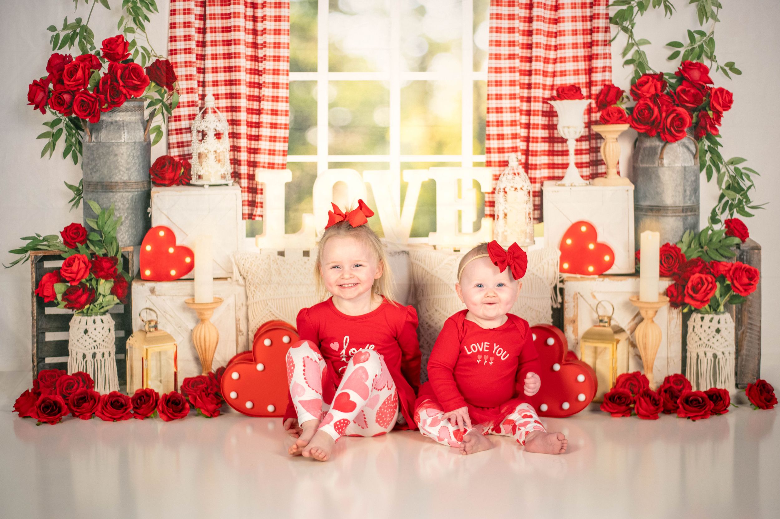 Two small children in red and white outfits sit among Valentine's Day decorations and roses.