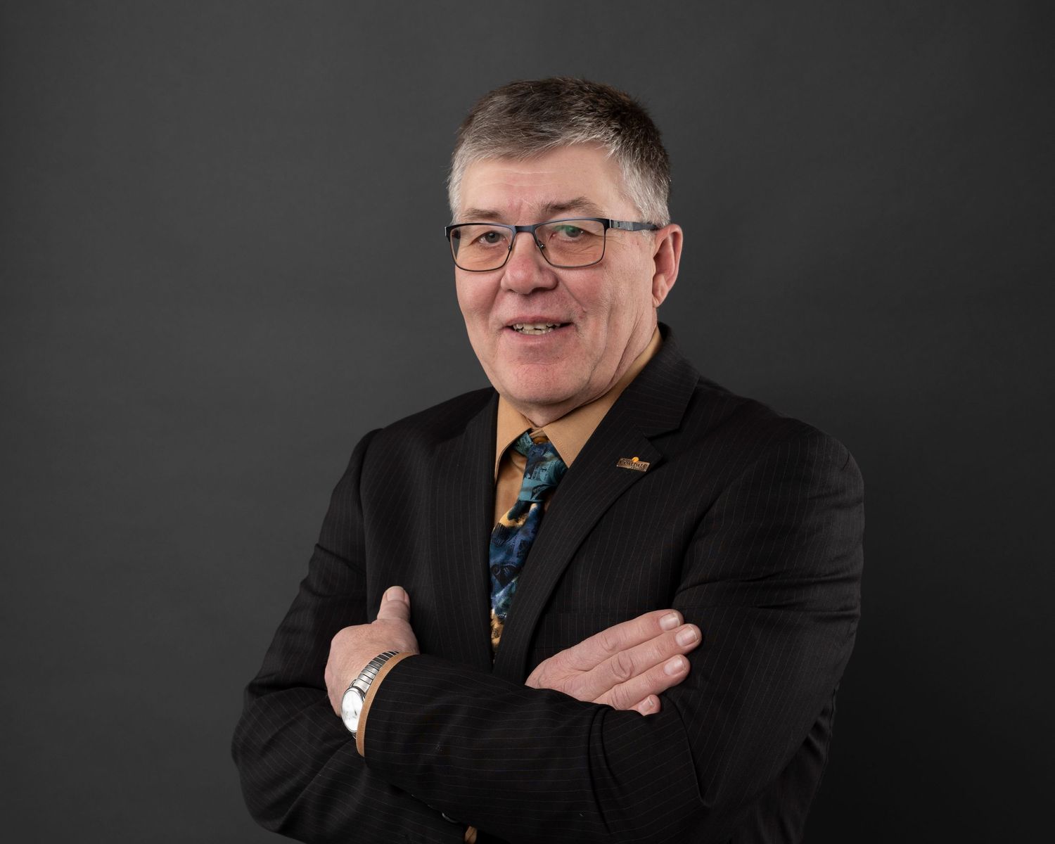 Business portrait of person in black suit with arms crossed wearing patterned tie against dark background.