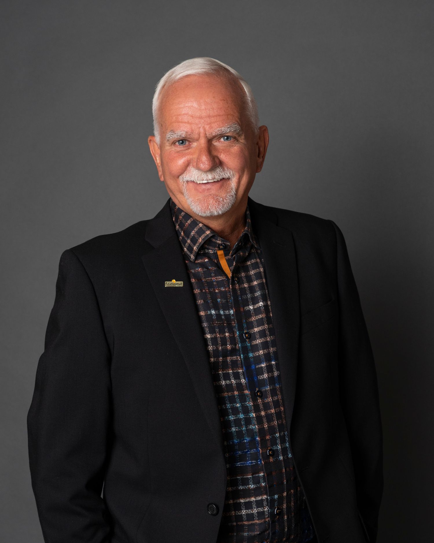 Professional portrait of person in black blazer and plaid shirt with white facial hair against dark background.