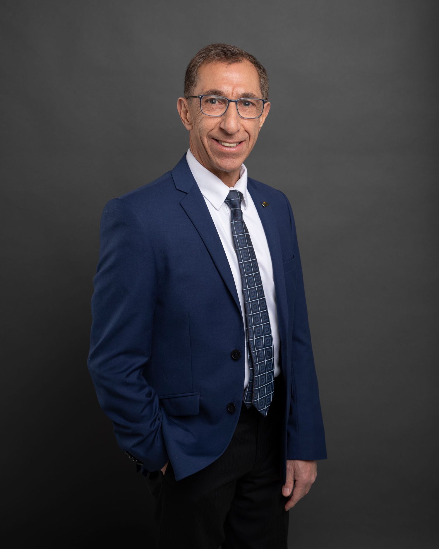 Professional headshot of person in navy suit and tie smiling against grey studio background.