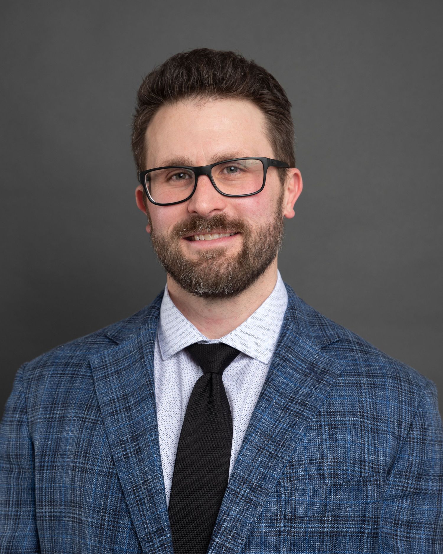 Professional headshot of person wearing glasses, a blue plaid blazer and black tie against grey background.