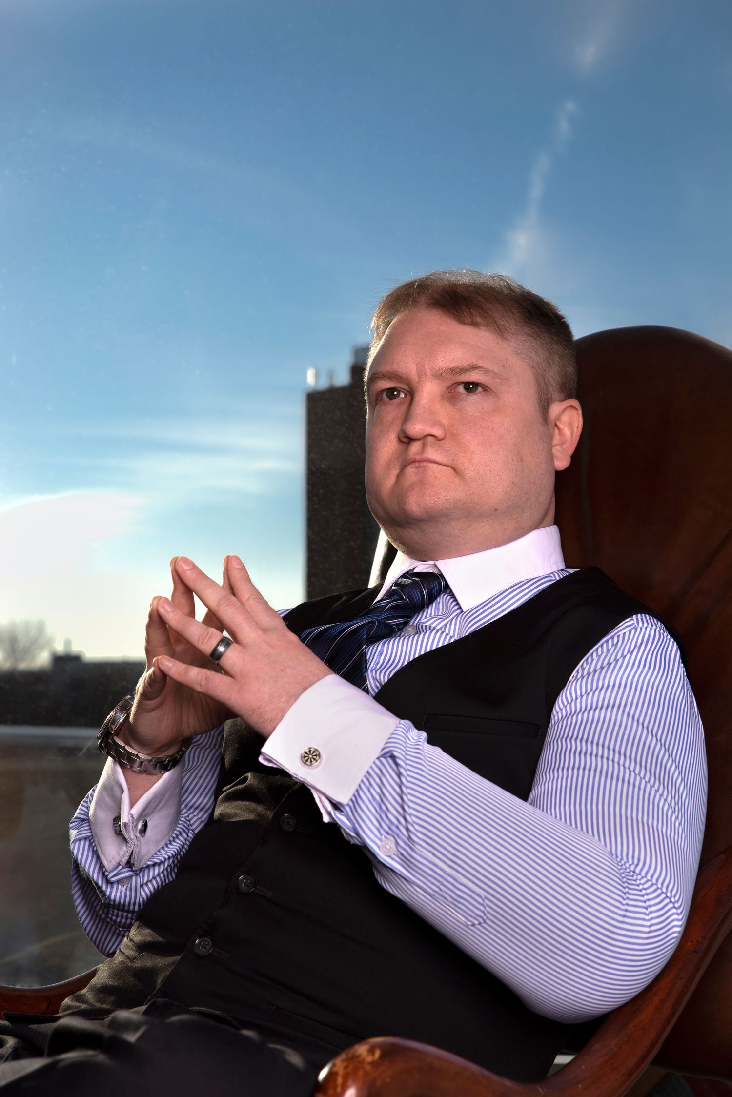 Person in business attire seated in leather chair with steepled fingers against bright sky background.
