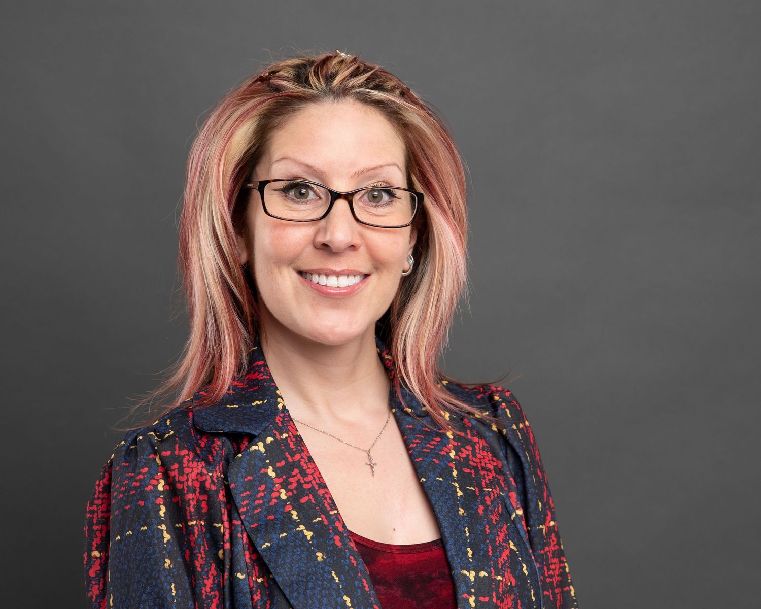 Professional portrait of someone with pink hair wearing glasses and patterned jacket against gray background.
