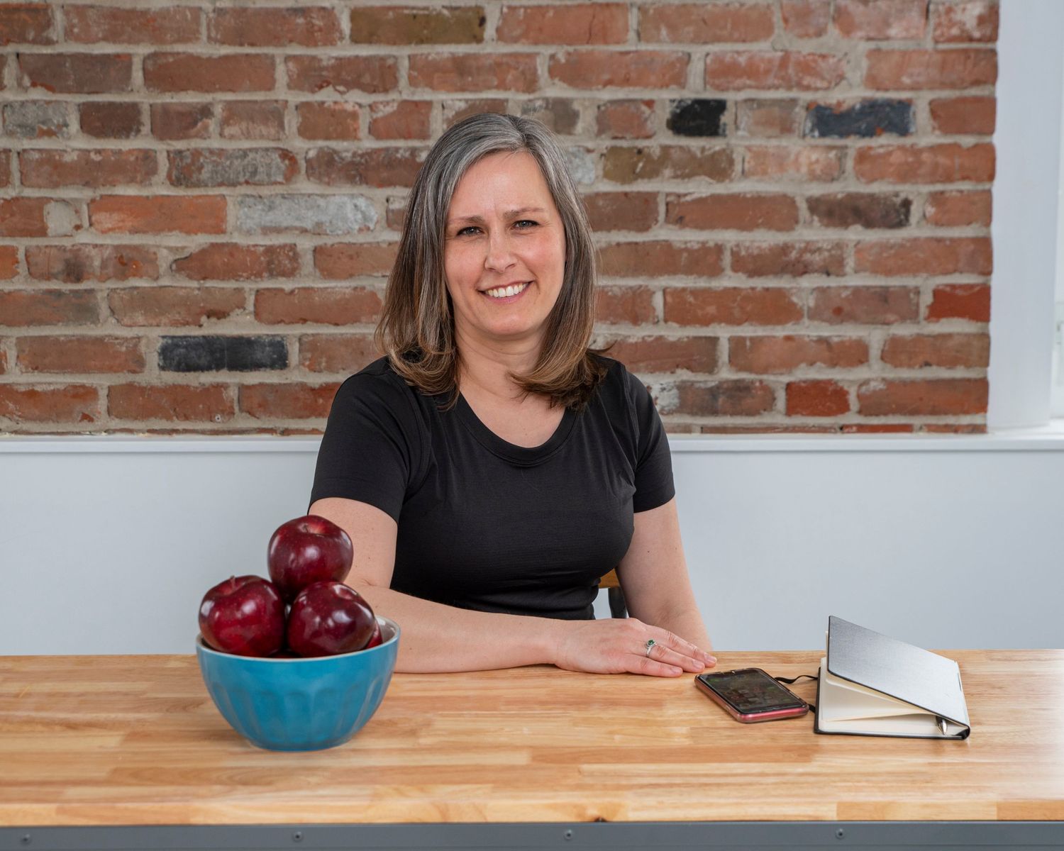 Woman at wooden table with bowl of red apples and notebook.